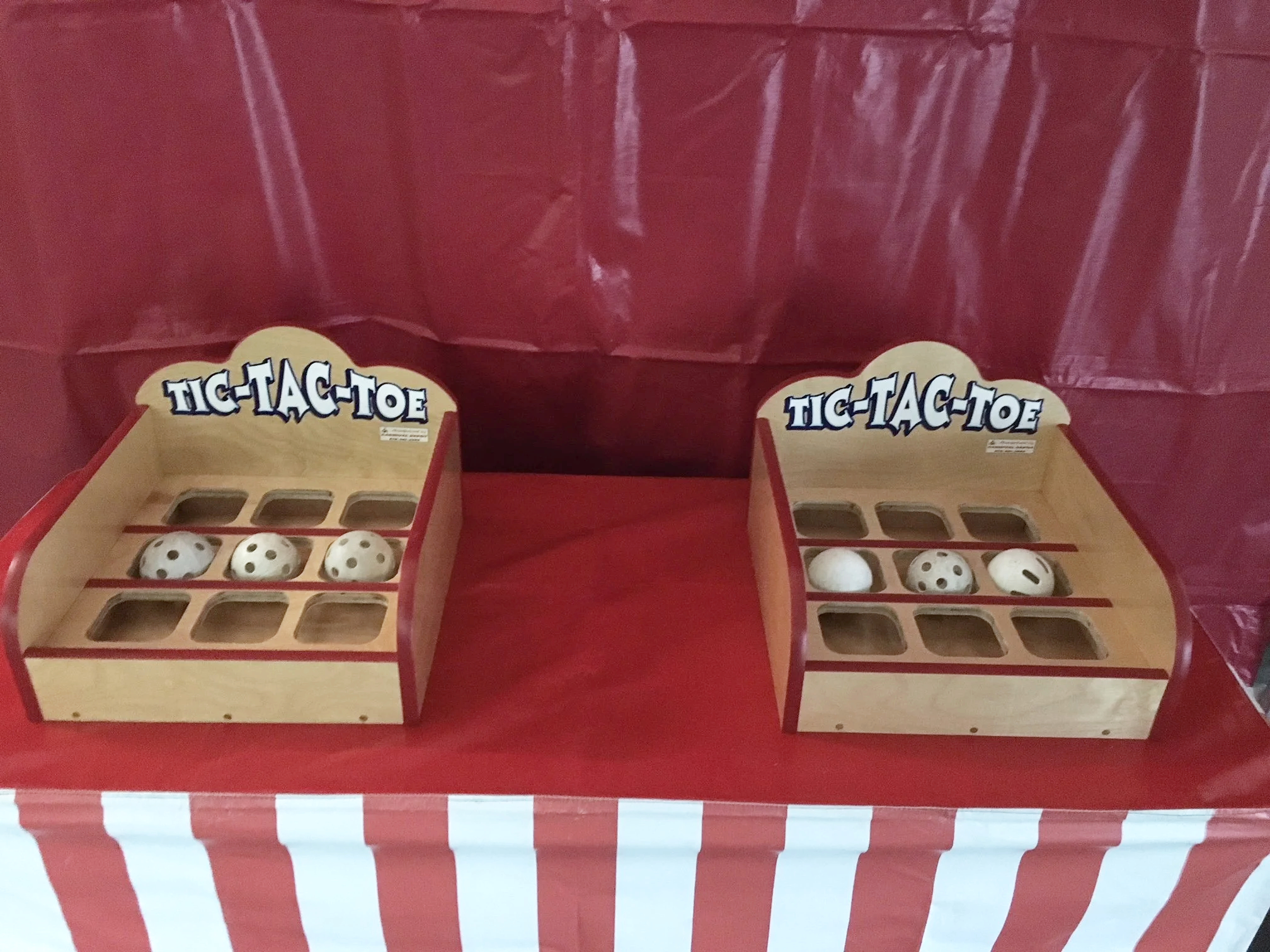A carnival-style game booth with two wooden targets labeled 'TIC-TAC-TOE' on each side, and four white balls with black dots inside the targets, set against a red-and-white striped background.