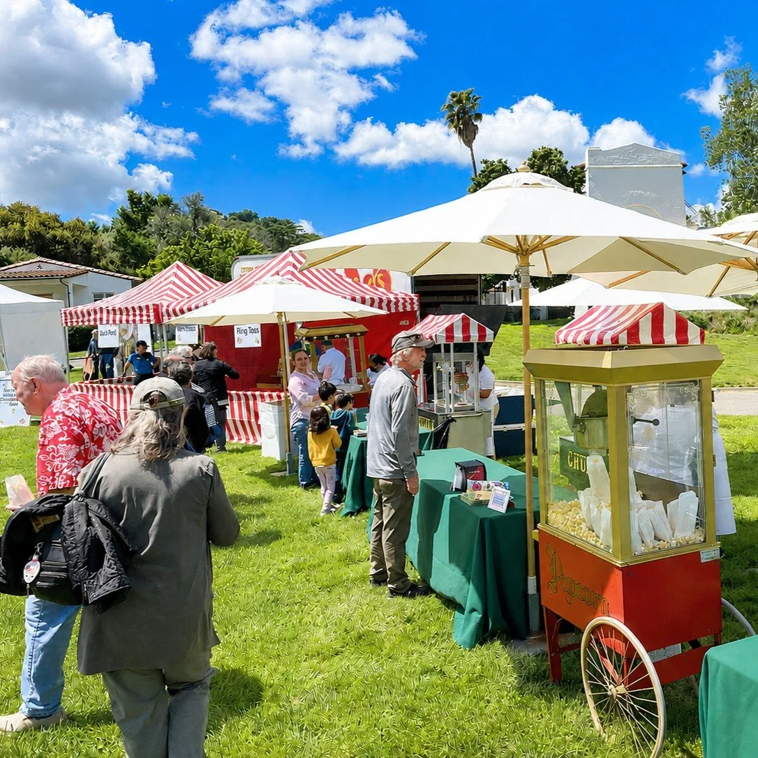 Outdoor fair with food booths, striped red and white umbrellas, and popcorn stand on green grass under a blue sky with clouds.