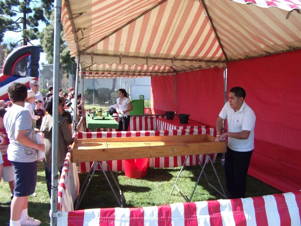 People attending a carnival game booth with a large wooden playing surface, a red and white striped tent, and onlookers in line playing the game outdoors on a sunny day.