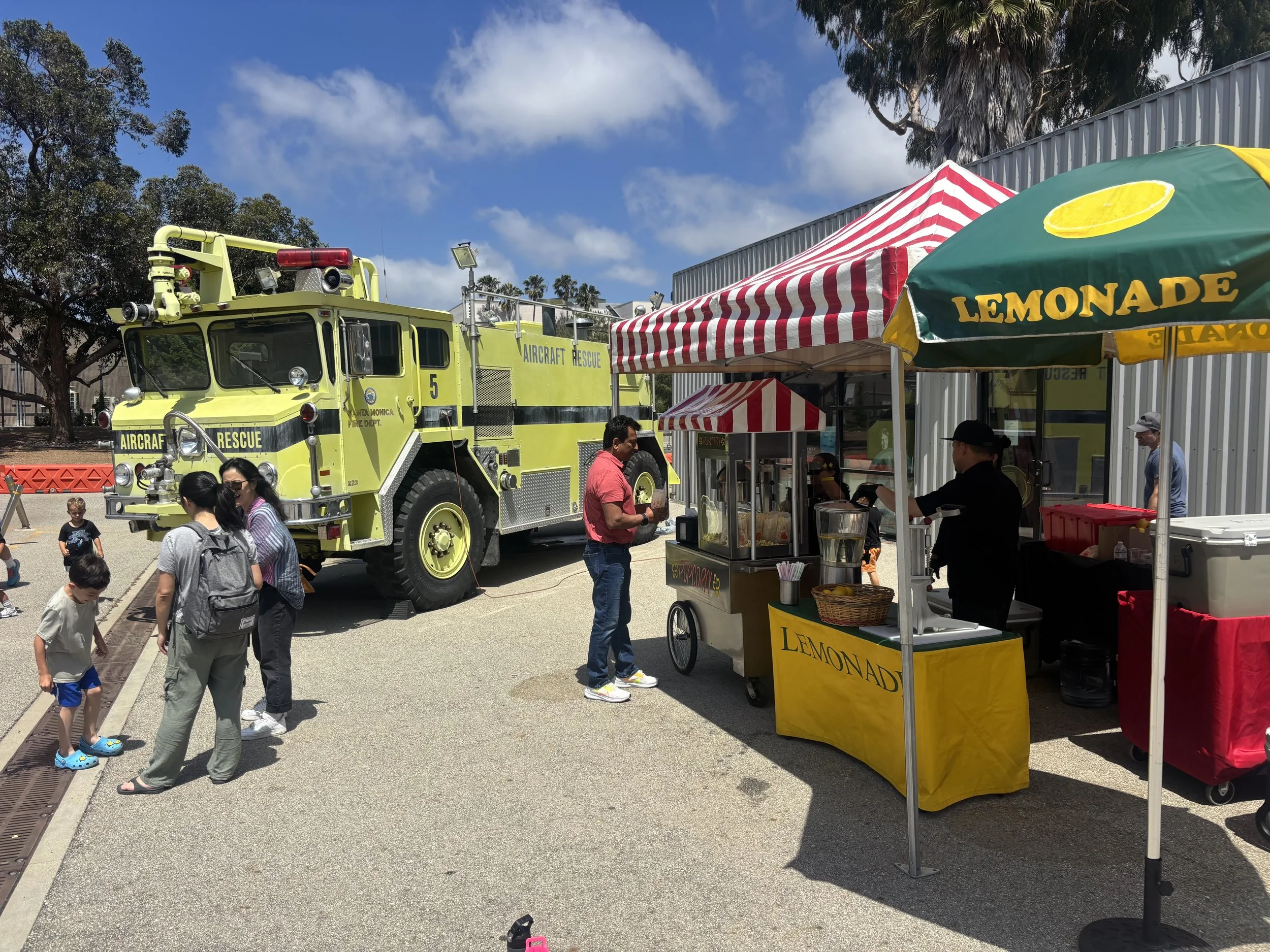 A yellow aircraft rescue fire truck is parked outdoors with several people and children nearby. There are tents, including a red and white striped tent and a yellow lemonade stand, under a partly cloudy sky.