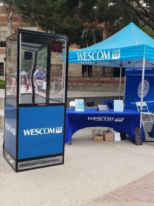 A Money Booth with a blue Wescom Financial Services table and tent, and a transparent phone booth in an outdoor setting. This is a photo of a "branded" Moneybooth