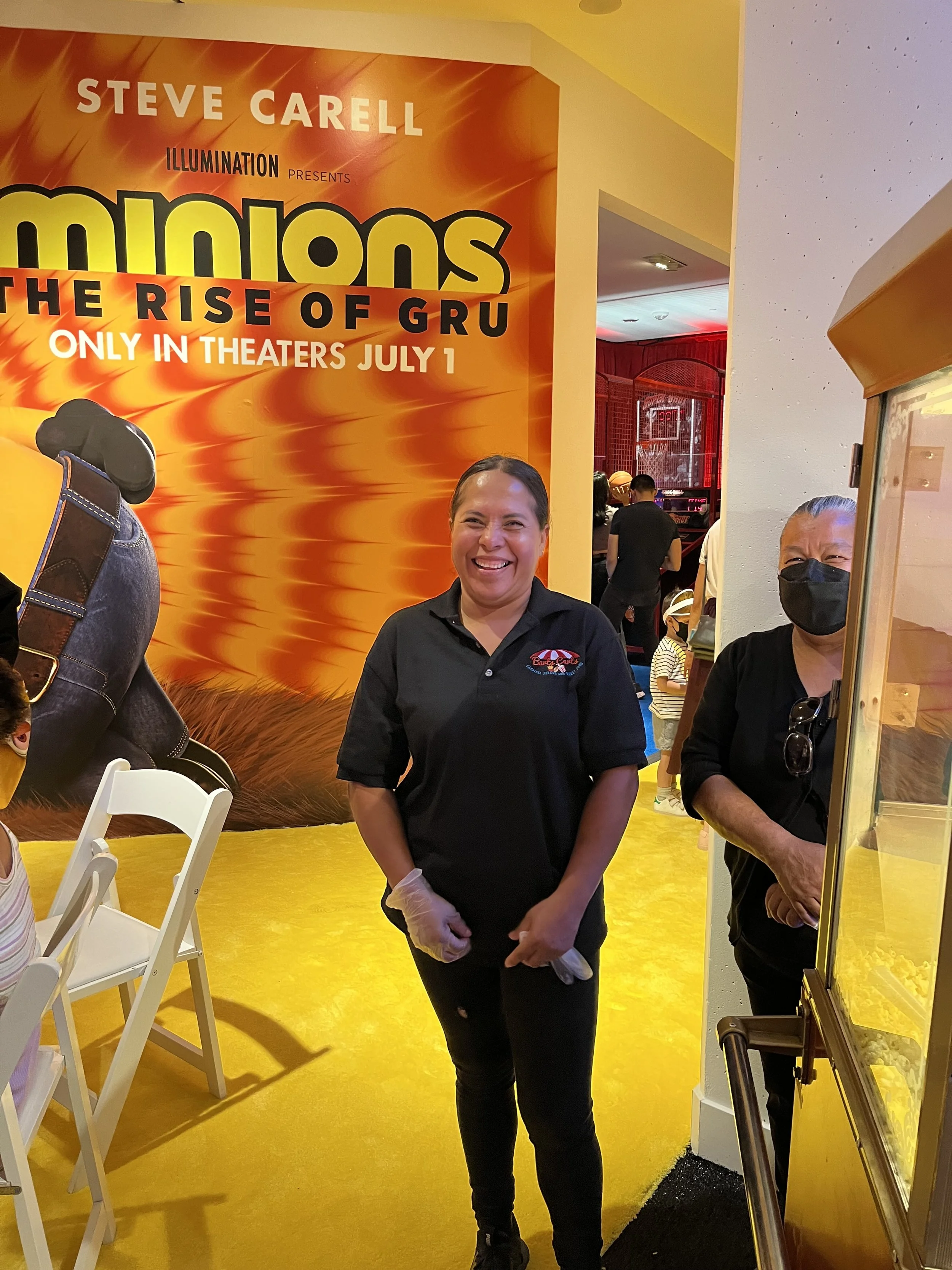A woman smiling in front of a poster for the movie 'Minions: The Rise of Gru' at a theater. She is wearing a black shirt with a logo and a glove on her left hand. Another person, wearing a black mask, is partially visible on the right.