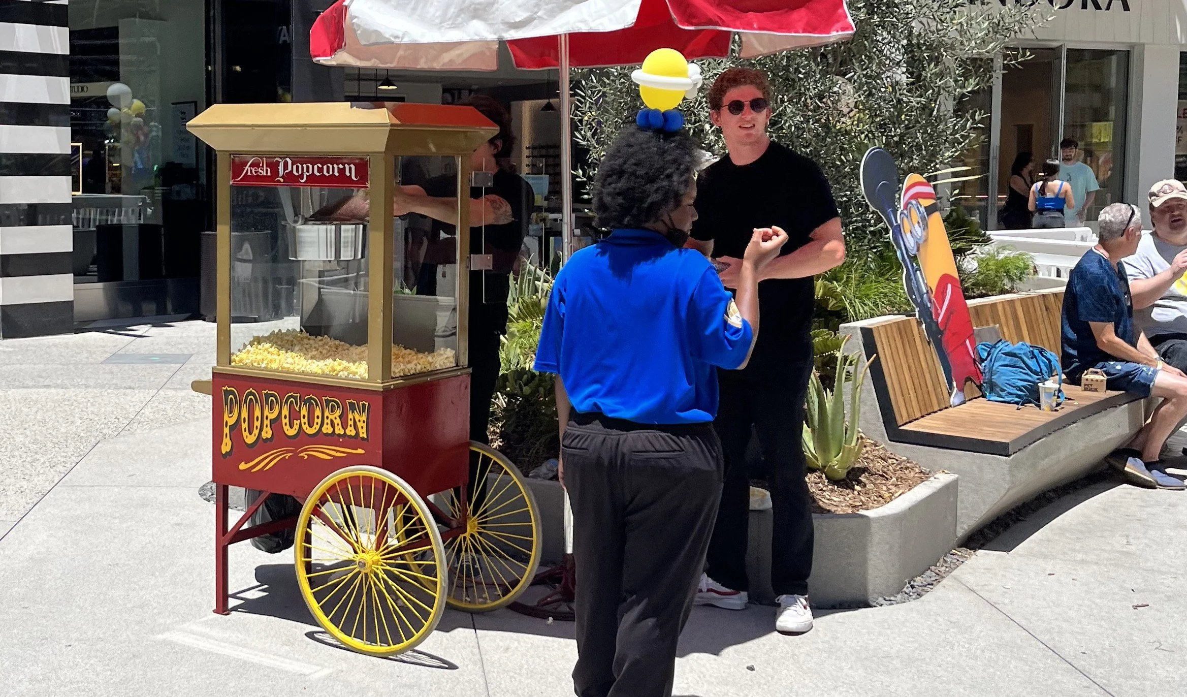 People standing near a popcorn cart on a city sidewalk, with a man wearing sunglasses and a black shirt talking to a woman in a blue shirt. Others are sitting on a bench and walking in the background.