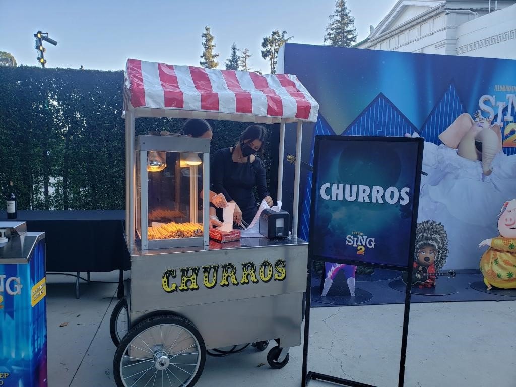 A churros stand at an outdoor event with a vendor preparing churros, a sign displaying the word "CHURROS," and decorative characters in the background.