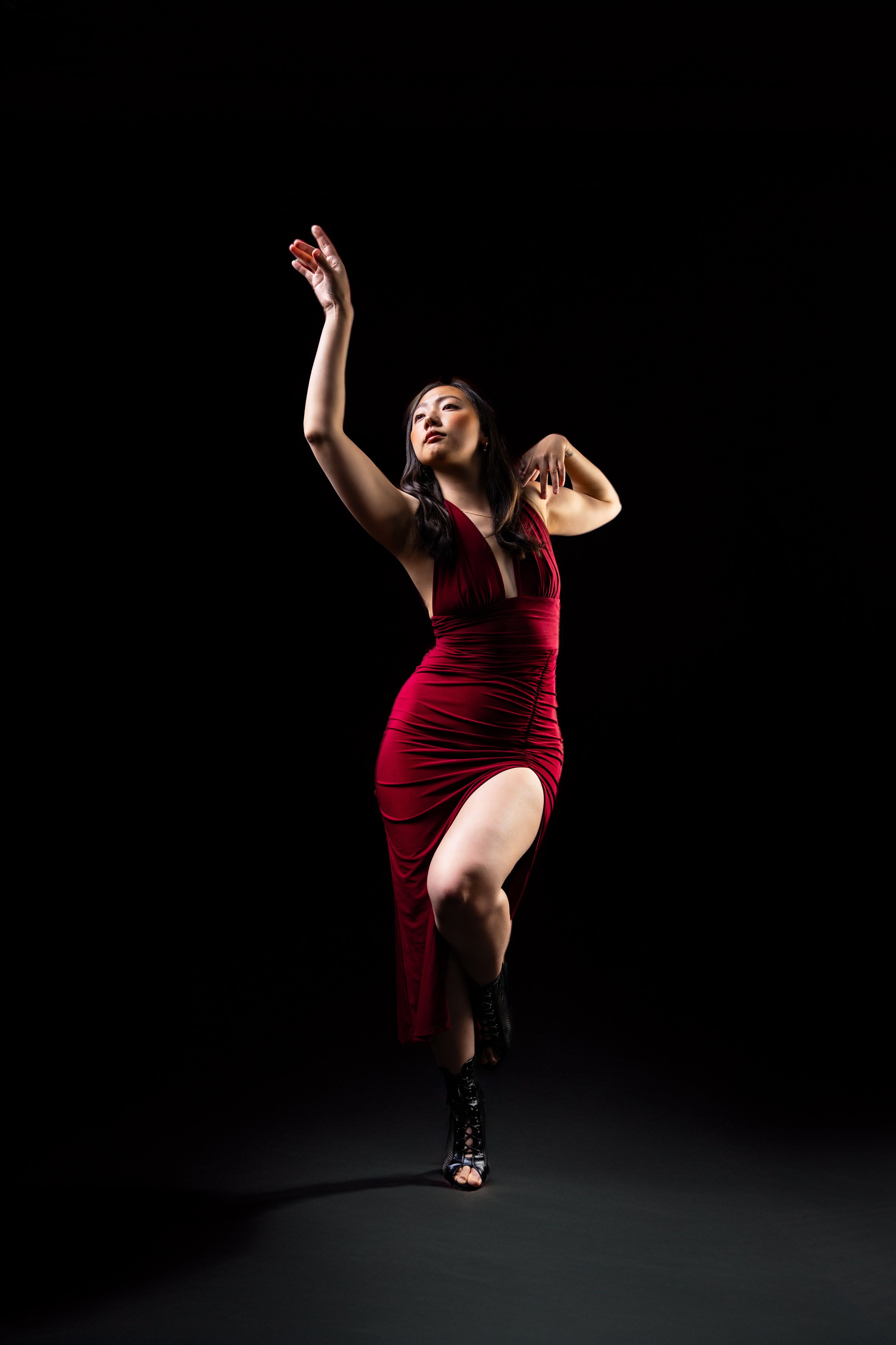 A dancer wearing a red dress and black high-heeled shoes poses against a black background, with one arm raised and the other bent near her face.