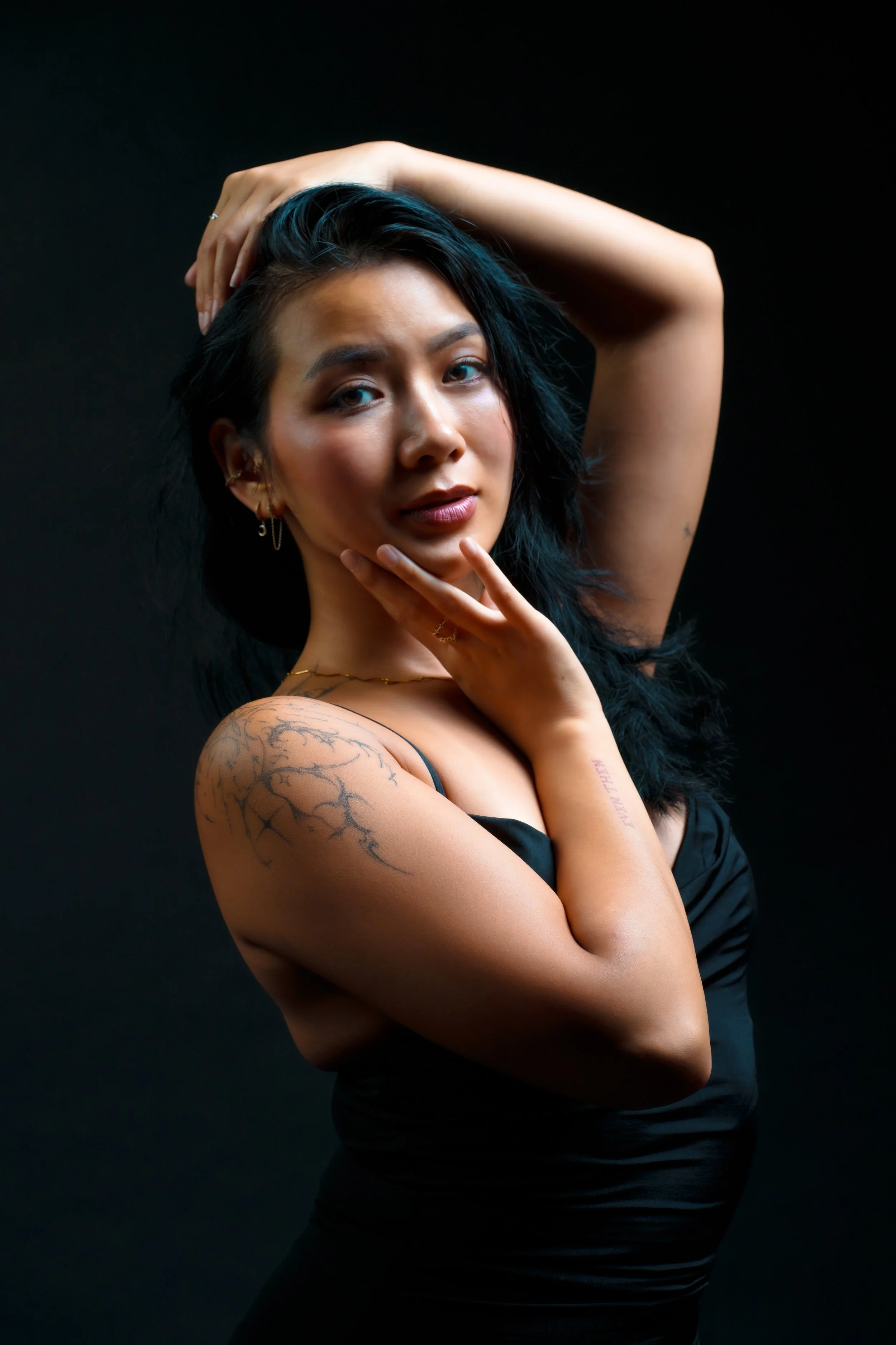 A young woman with black hair, tattoos on her arm, and earrings, is posing against a black background with her hand on her chin and the other arm raised behind her head.