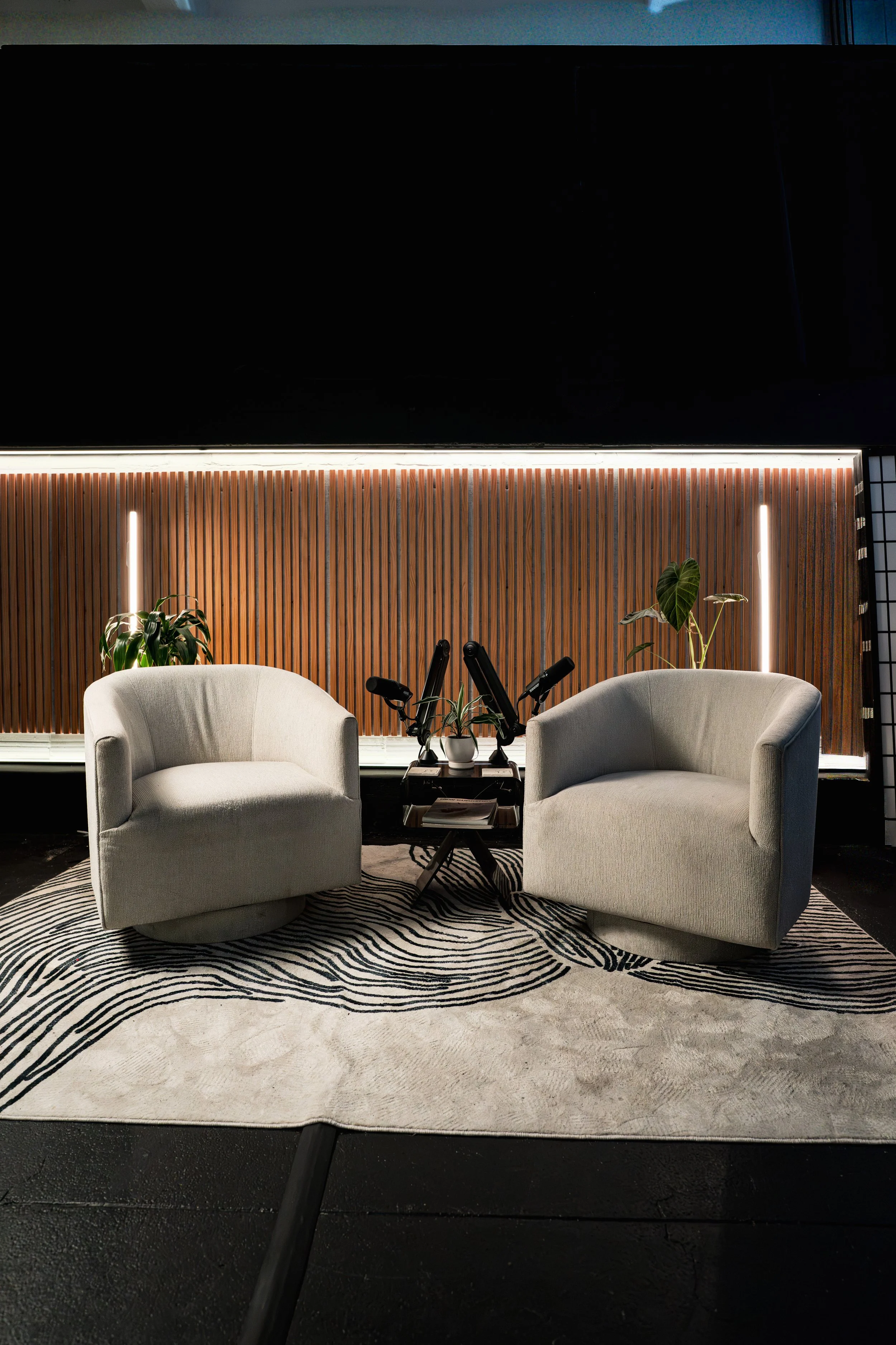 A modern interior space featuring two light grey armchairs placed on a black and white zebra-patterned rug, two potted plants, a small table with decorative items, a wooden slat wall with LED strip lighting, and a dark upper wall with a window.