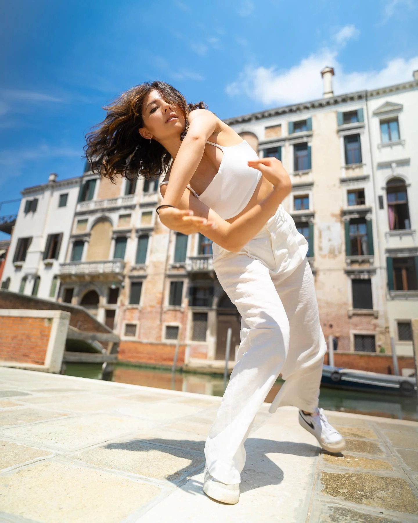 Woman performing dance move outdoors on paved surface with old building in background, wearing white pants, a white tank top, and sneakers.