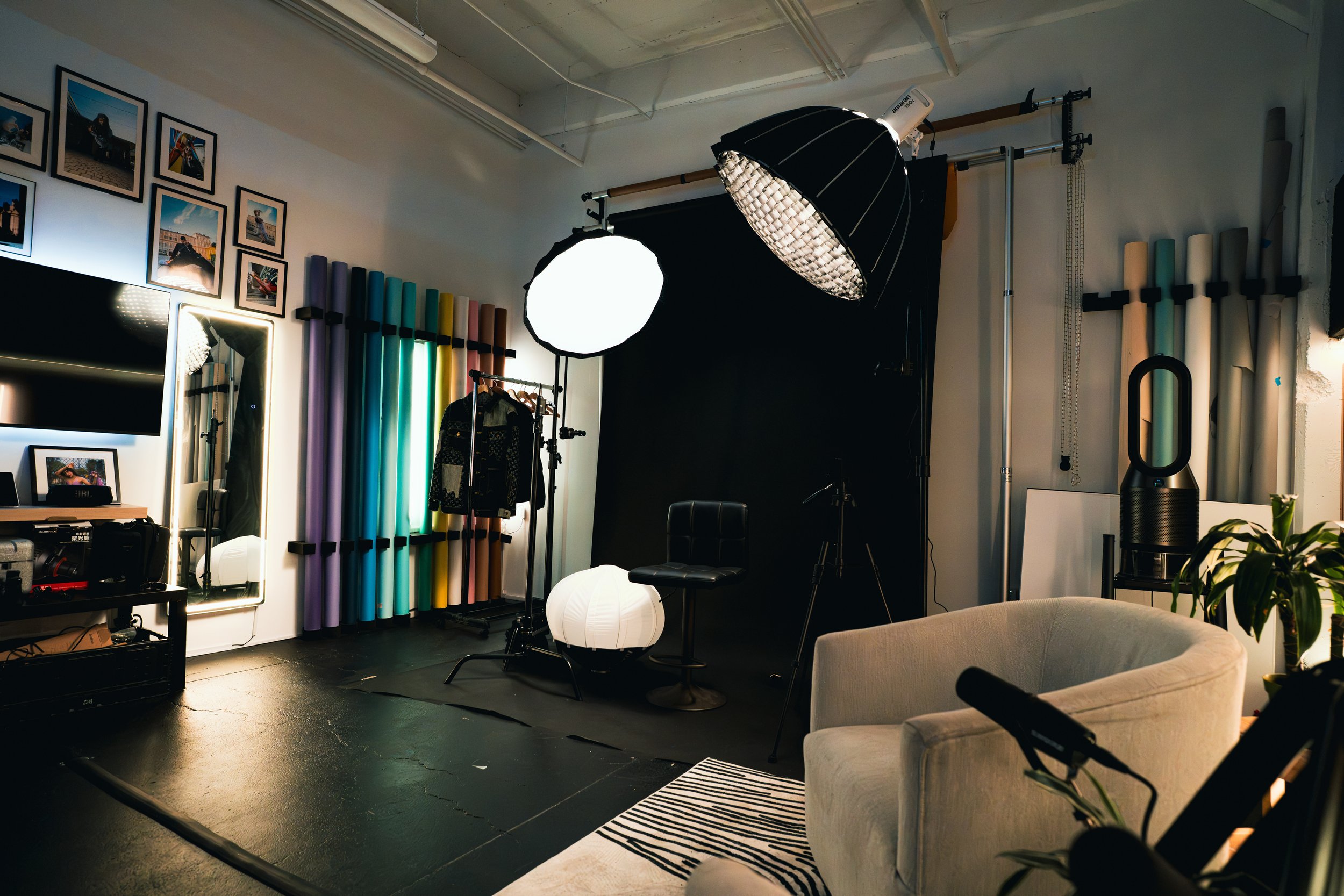 Photography studio with professional lighting equipment, a black backdrop, colorful rolled paper backgrounds, a mirror, framed photos on the wall, a computer monitor, and furniture.