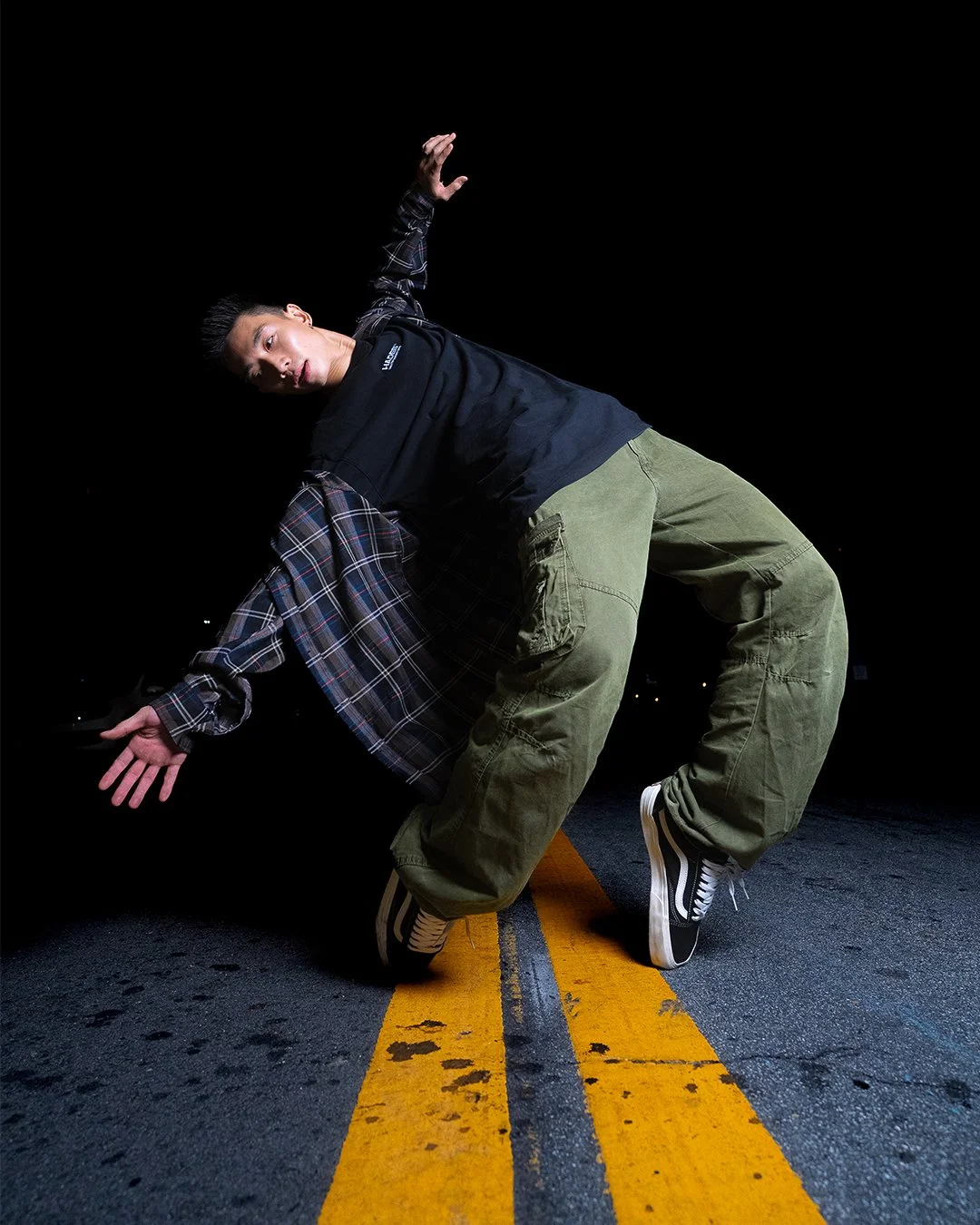 A young man posed on a dark street at night, balancing on one hand and foot, with the other hand extended, against a black background and a yellow street line.
