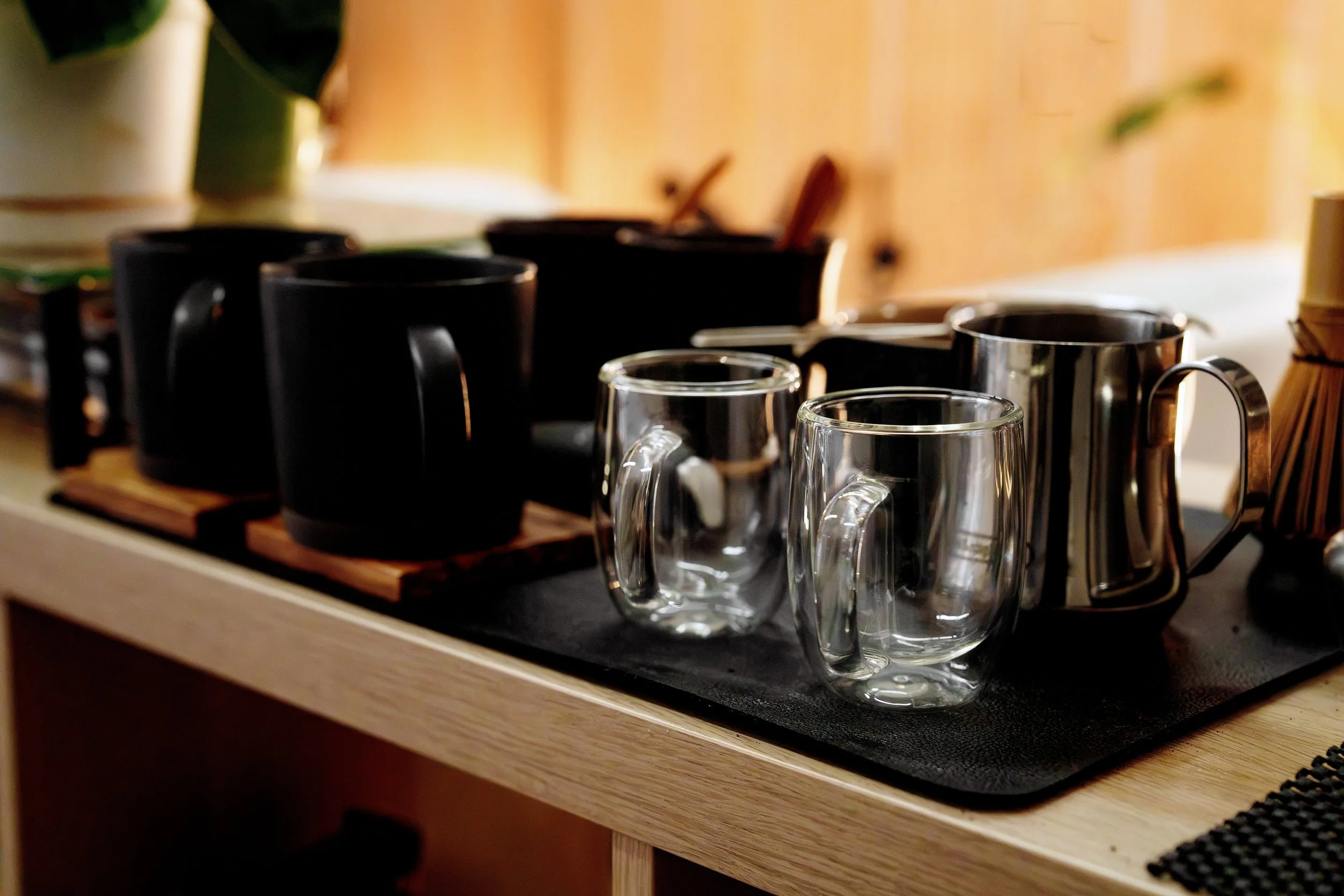 A collection of black mugs and clear glass coffee mugs on a wooden tray, with a plant in the background.