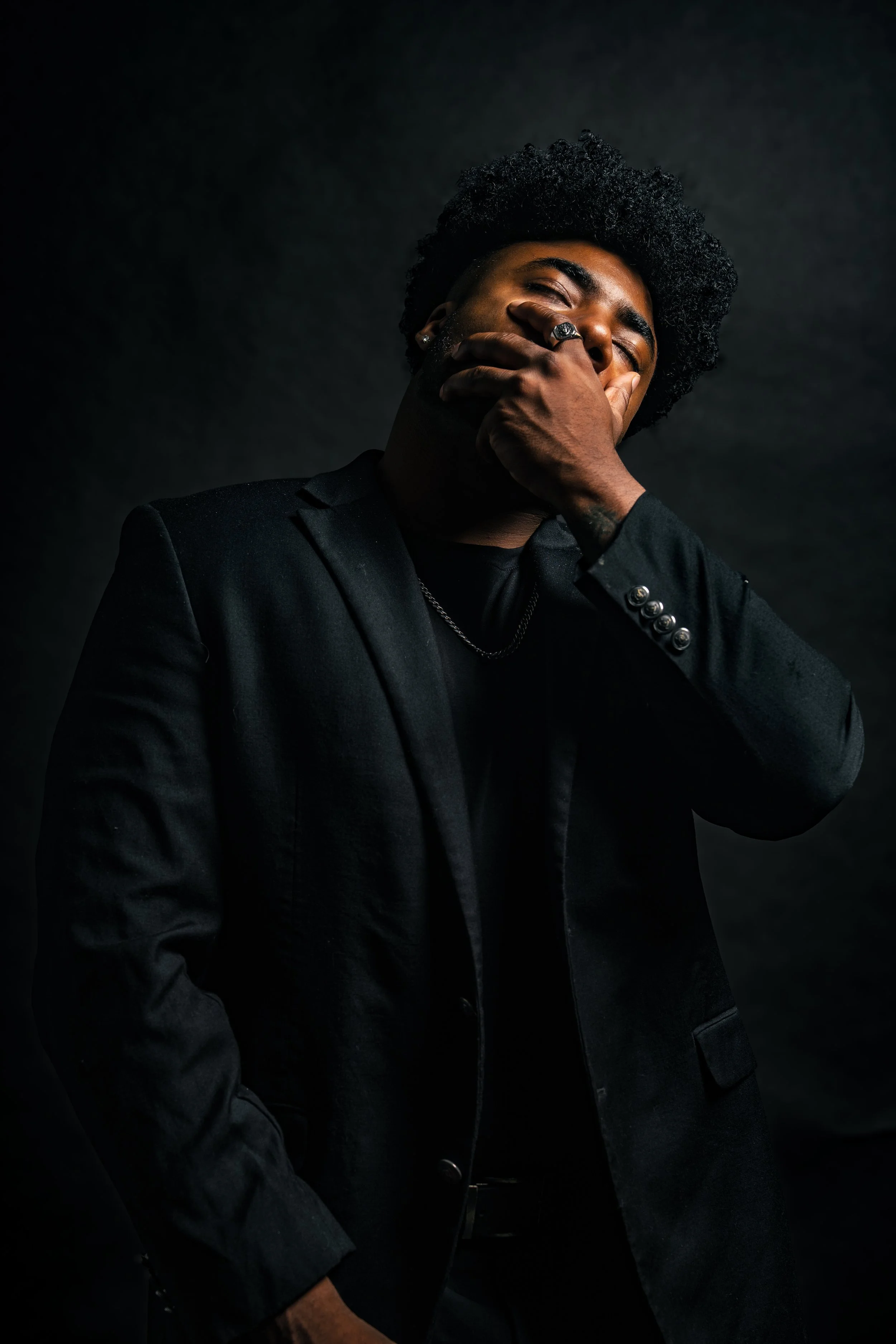 A man with curly black hair and dark skin wearing a black suit with silver button details, a black shirt, and a chain necklace. He covers his mouth with his right hand, wearing rings, and is standing against a dark background.