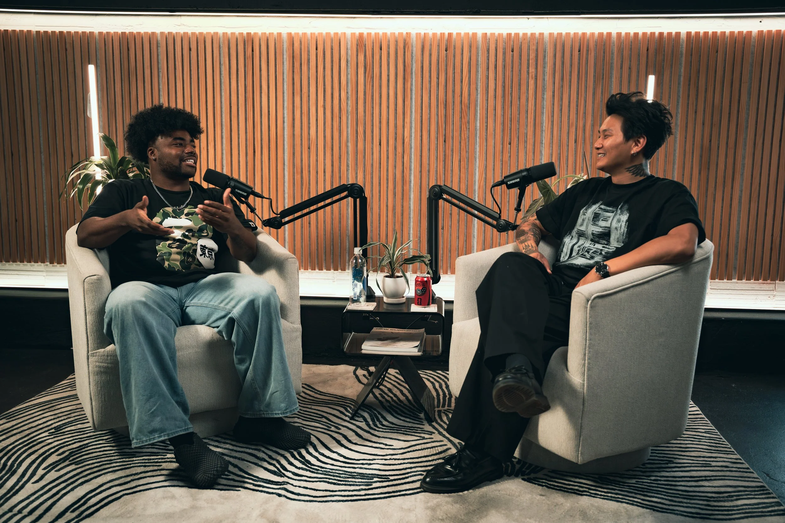 Two men having a conversation in a podcast studio. They are sitting in armchairs with microphones, with a wooden slat wall and plants in the background.