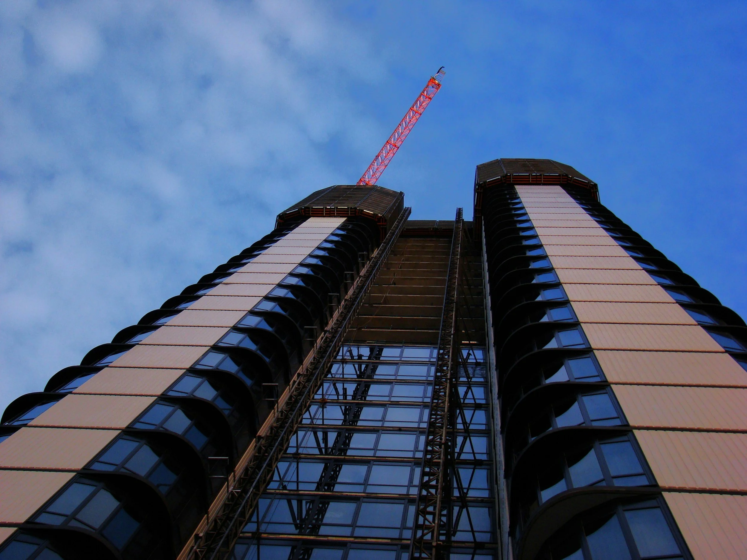 A high-rise building under construction with a red construction crane at the top, set against a partly cloudy blue sky.