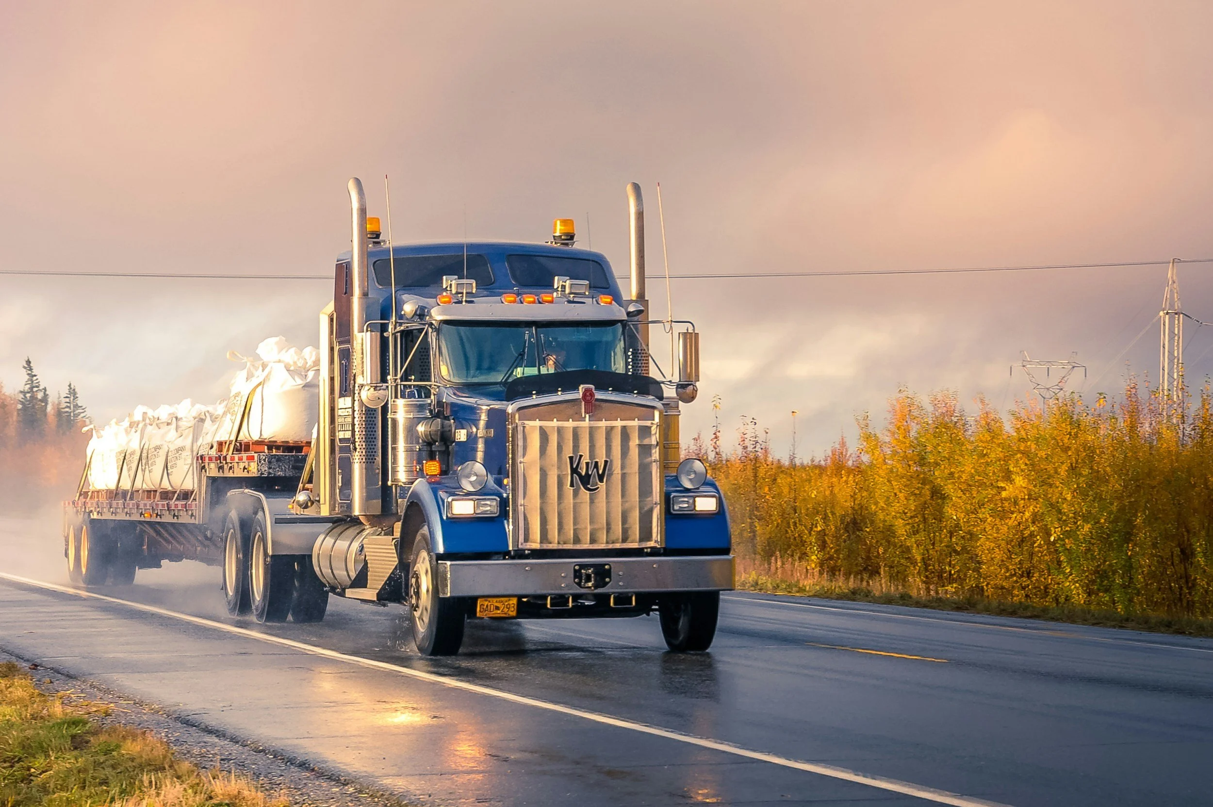 A blue semi-truck driving on a wet road during sunset, with a farm or rural area with trees and electrical poles in the background.