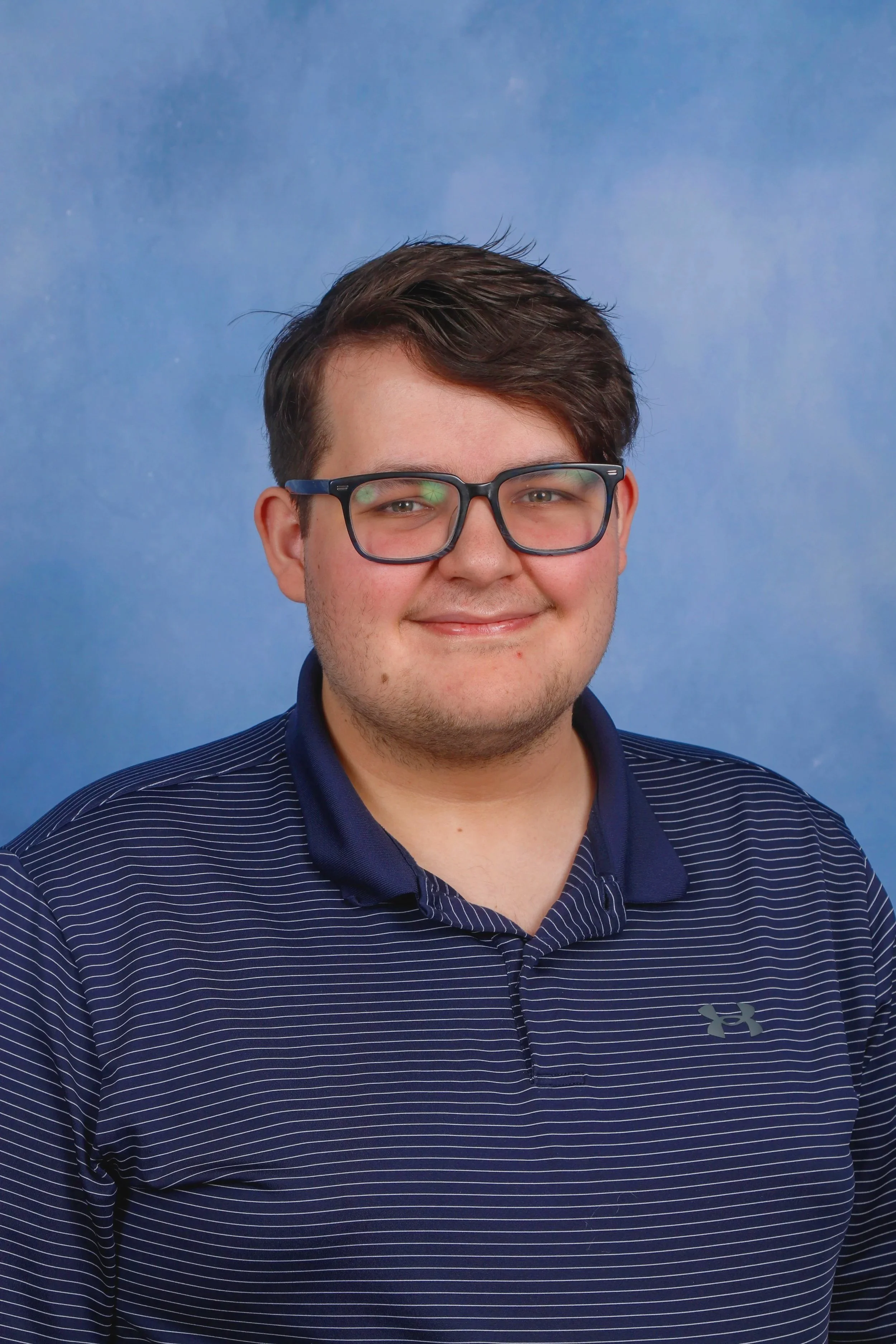 A young man with dark hair and glasses smiling wearing a navy blue Under Armour polo shirt with thin white stripes, standing against a blue background.