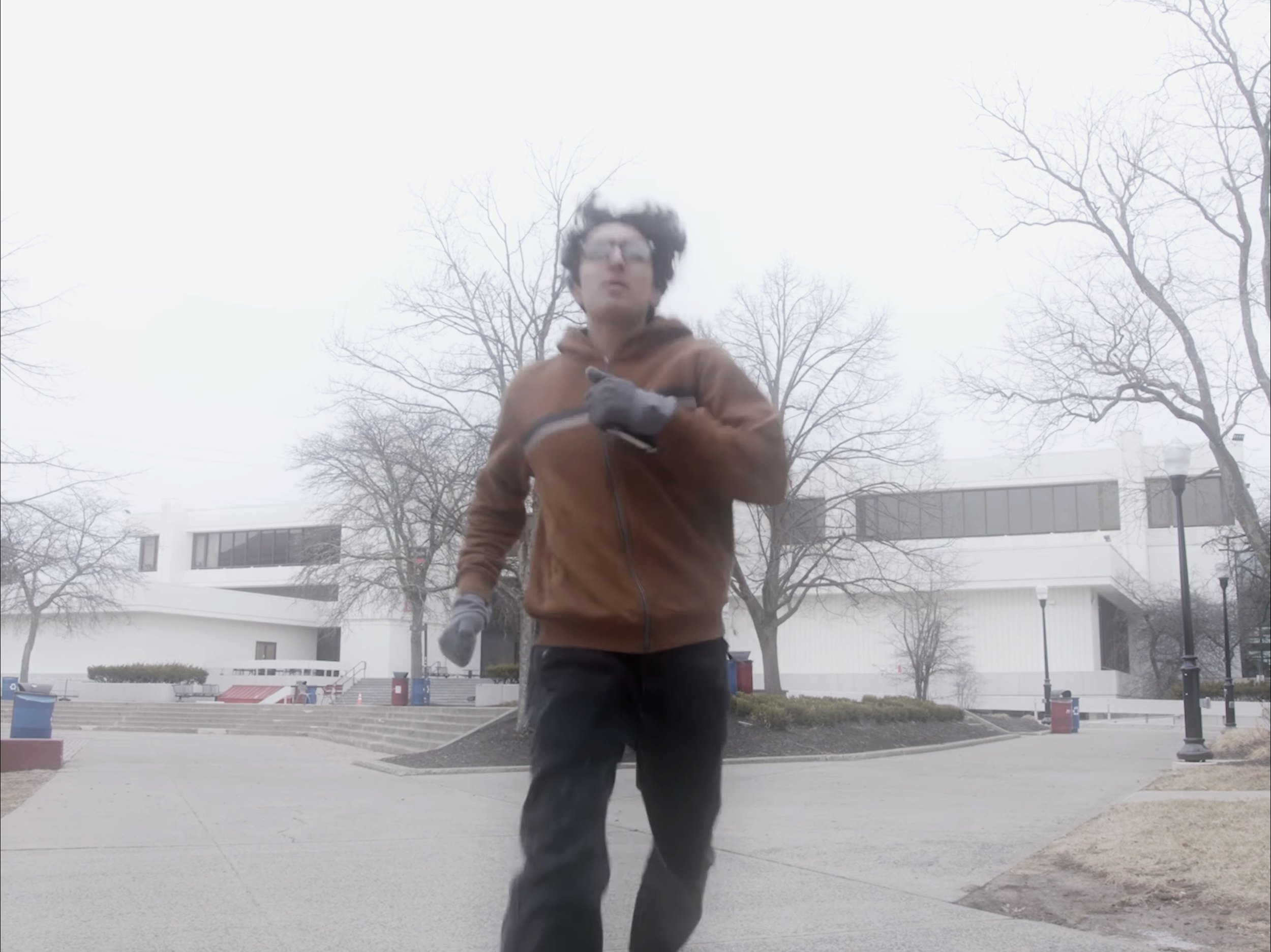 A person with curly dark hair and glasses running outdoors on a foggy day, wearing a brown jacket and gray gloves, with leafless trees and modern buildings in the background.