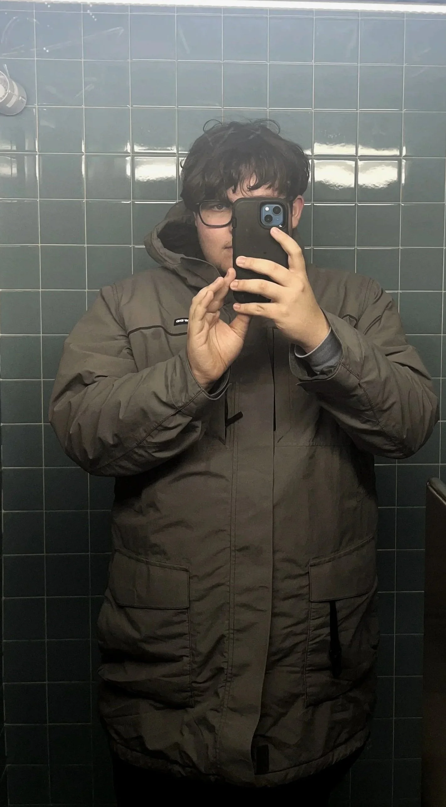 A person taking a mirror selfie in a bathroom with dark gray tiled walls, wearing glasses and a brown jacket.