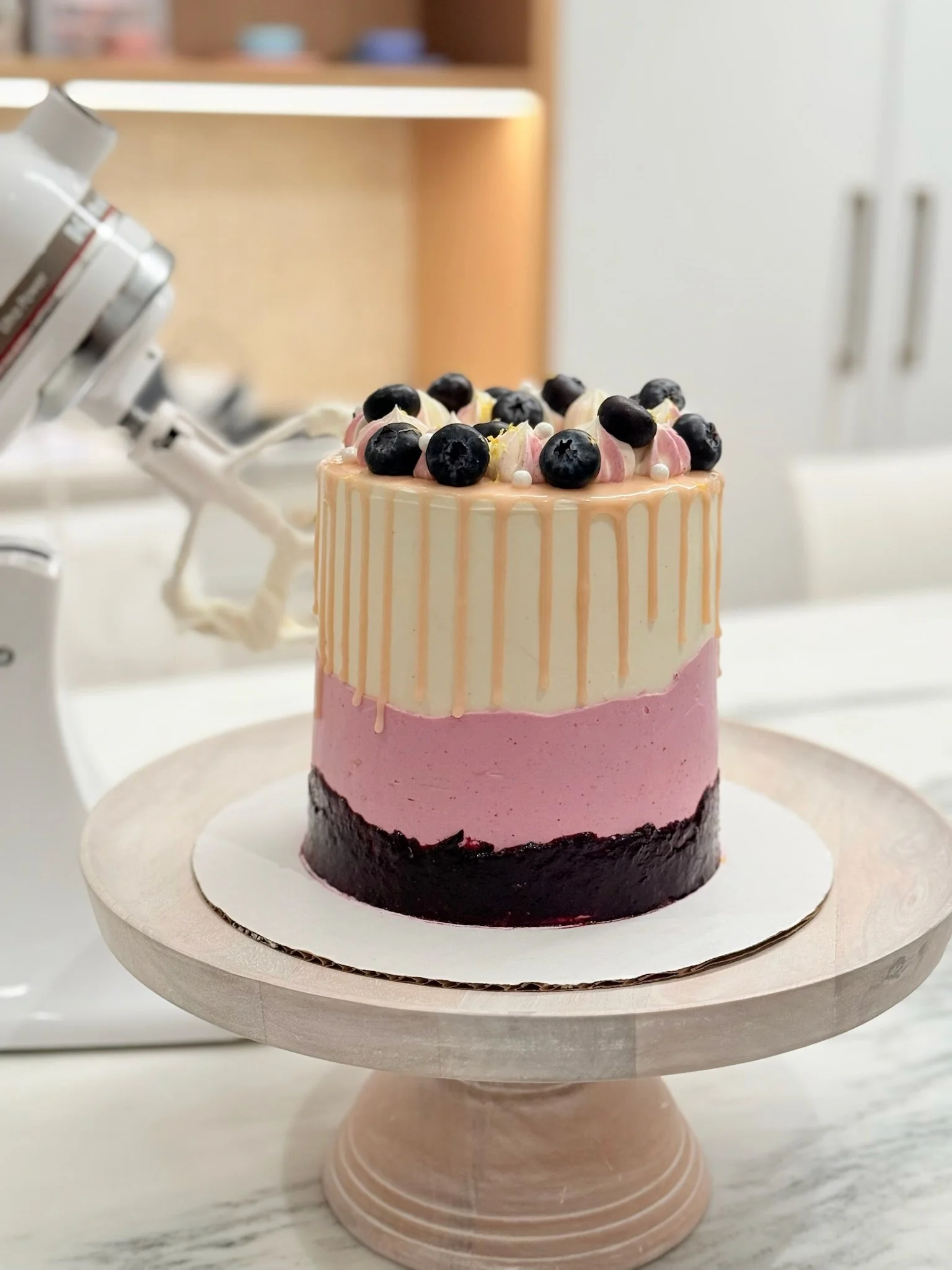 A tiered cake with pink and white layers, topped with blueberries and decorative cream swirls, on a cake stand.