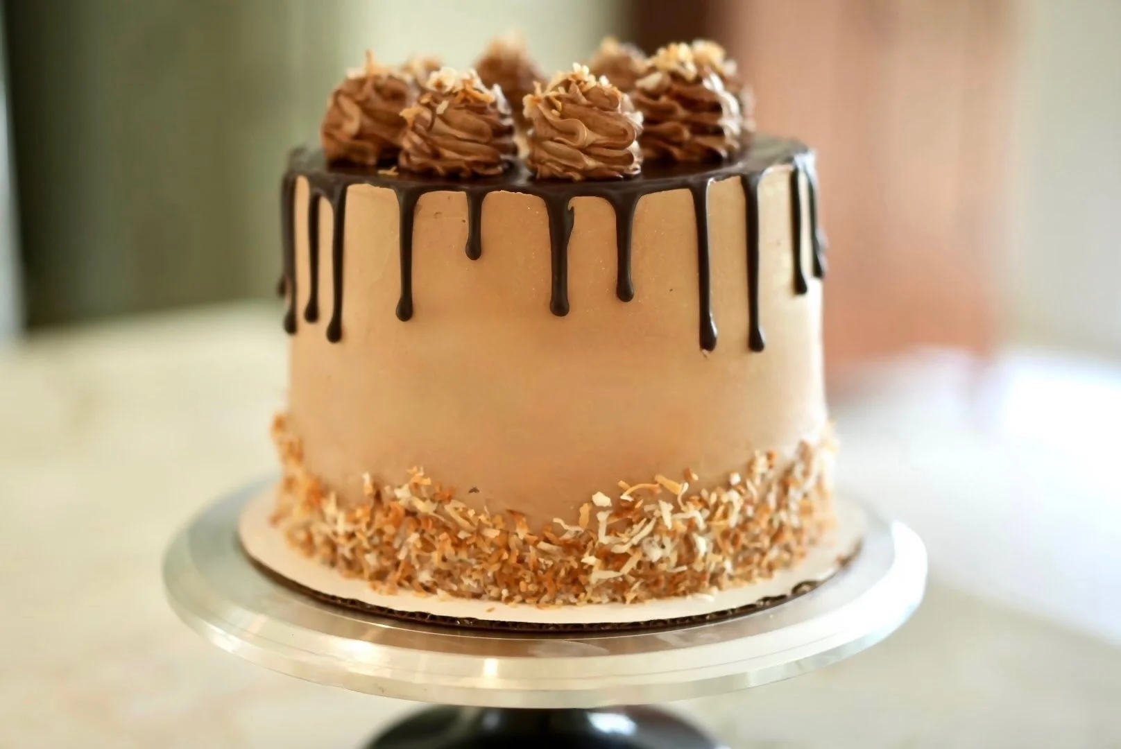 A tall birthday cake with chocolate drip icing, topped with whipped cream rosettes, and decorated with toasted coconut around the base.