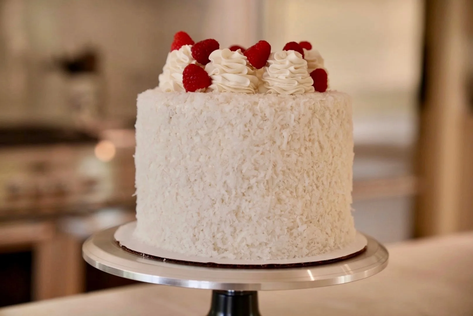 White rectangular cake with shredded coconut exterior, topped with whipped cream swirls and fresh raspberries, on a metallic cake stand.