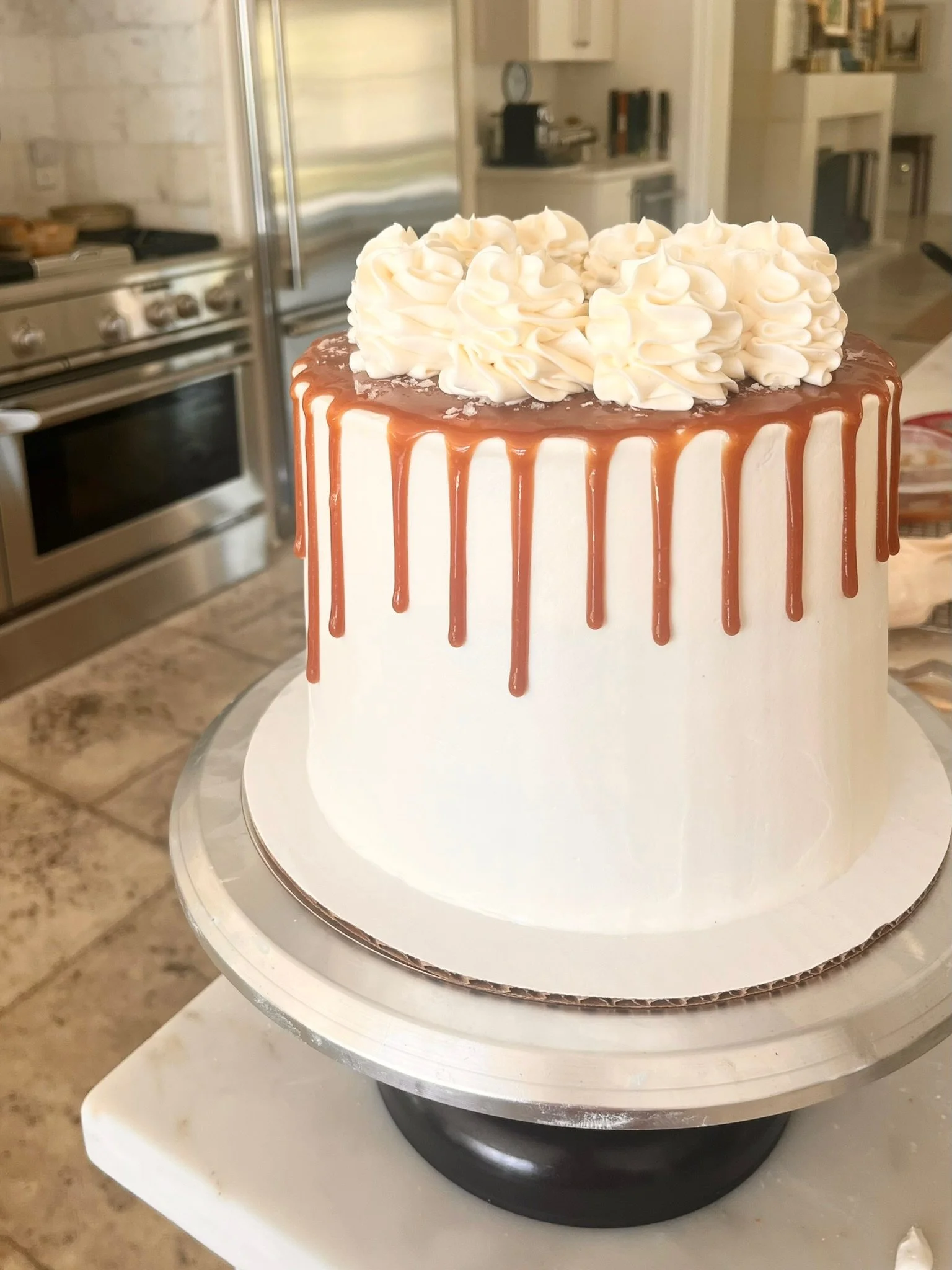 A tall white frosted cake with caramel drip icing and white piped frosting on top, placed on a white cake stand in a kitchen.