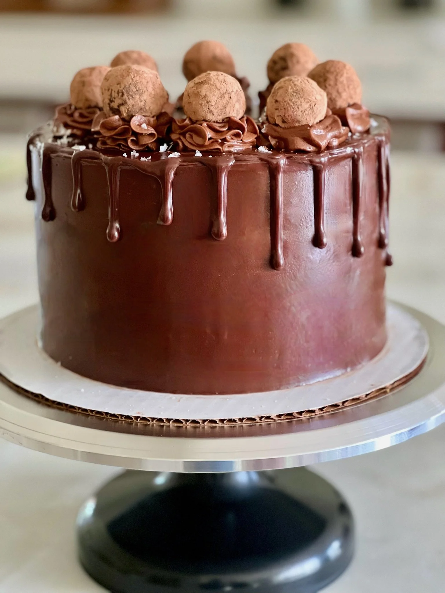 A round chocolate cake decorated with chocolate frosting, chocolate truffles on top, and a chocolate drip glaze on the sides, on a cake stand.