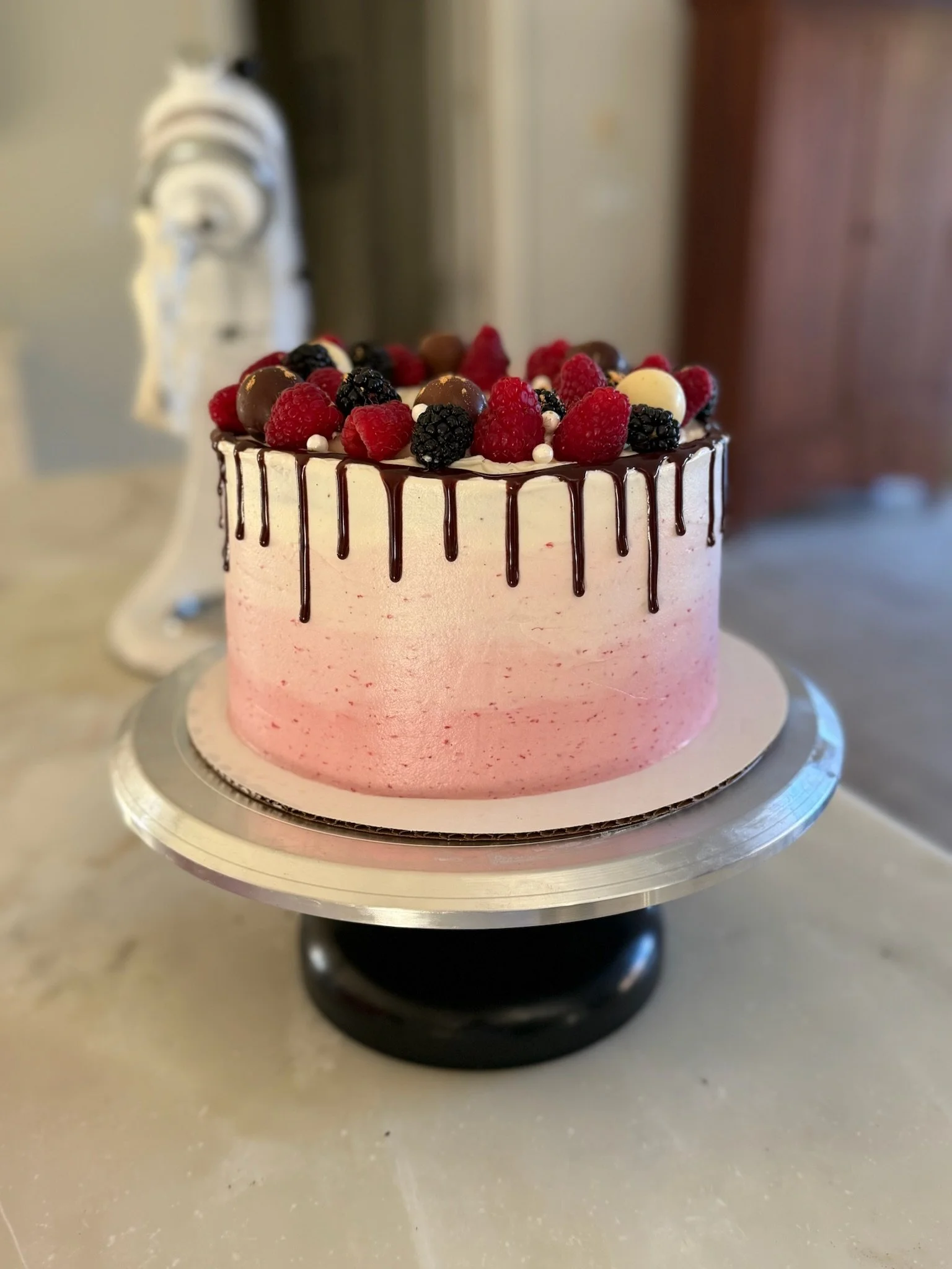 Close-up of a pink layered cake decorated with berries, white and brown chocolates, and a chocolate drizzle on top, sitting on a silver cake board.