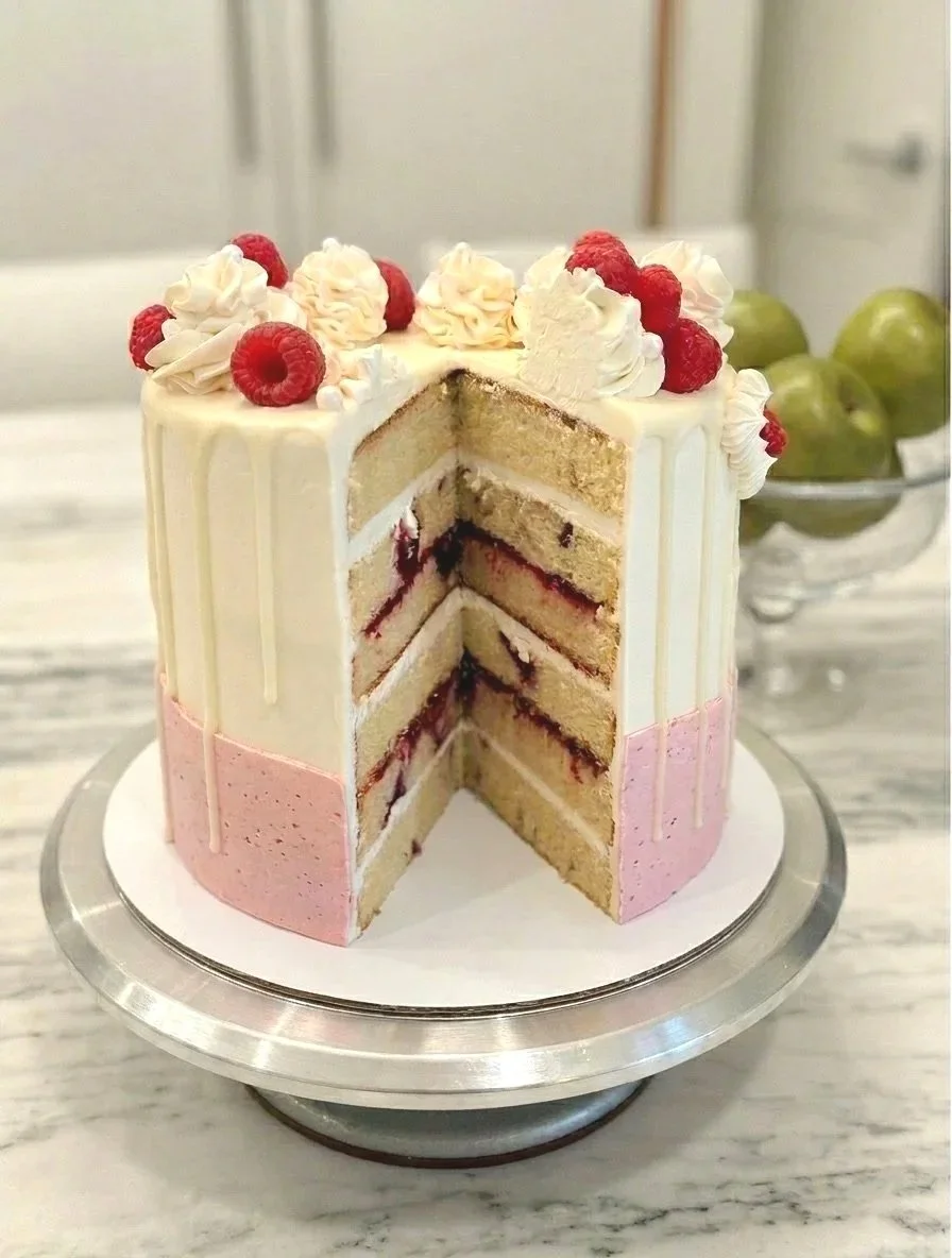 A multi-layered cake with white and pink frosting, topped with whipped cream and raspberries, with a slice removed showing layers of vanilla cake, berry filling, and cream.