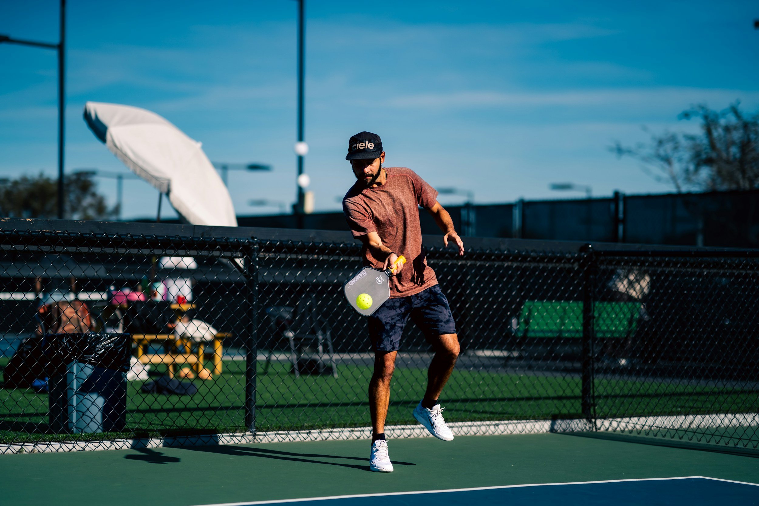 A man playing pickleball on an outdoor court during daytime, wearing a black cap, brown T-shirt, and dark shorts, holding a paddle and hitting a yellow pickleball.