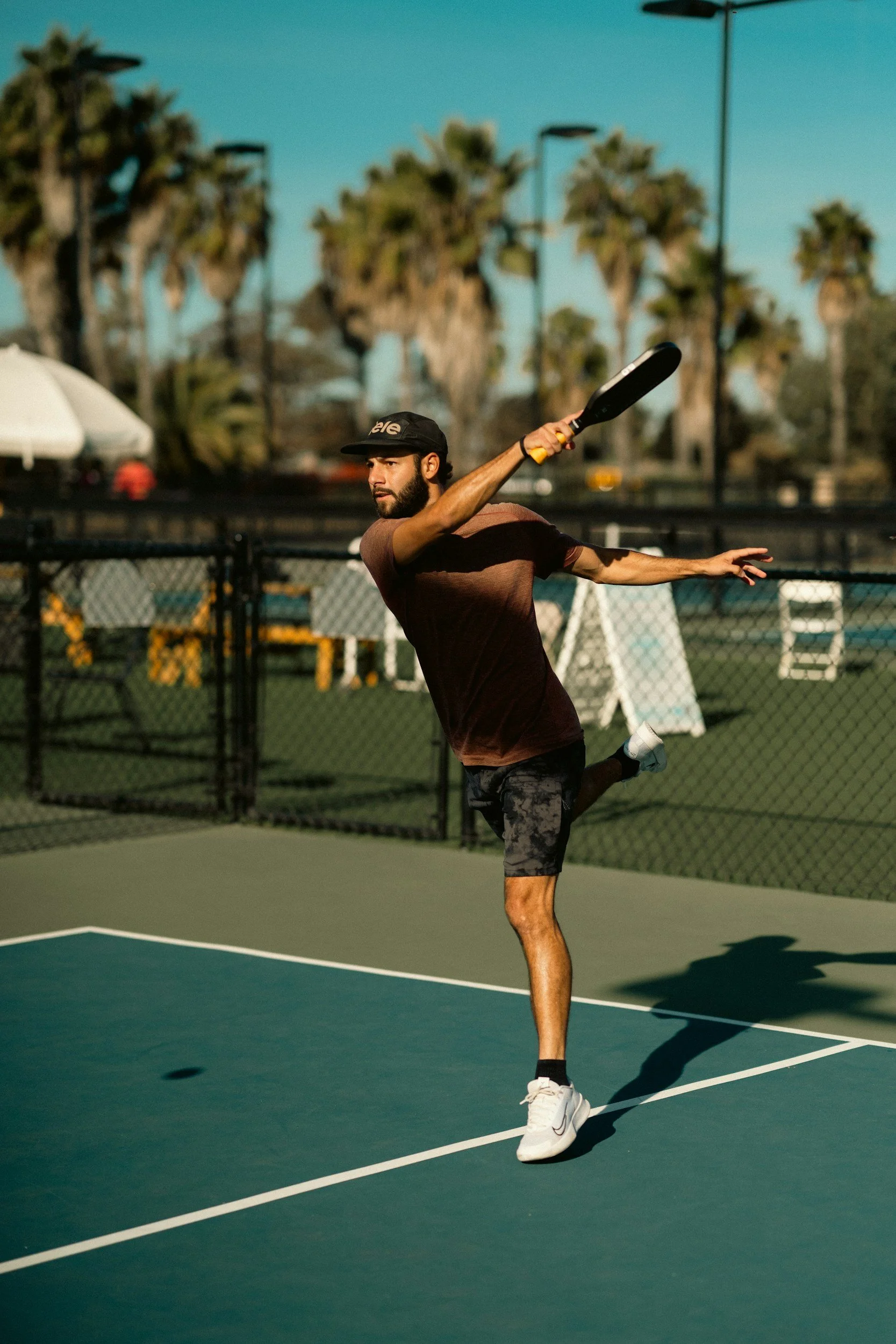 A man playing tennis on an outdoor court, wearing a black cap, brown shirt, black patterned shorts, white shoes, and holding a tennis racket, with trees and street lamps in the background.