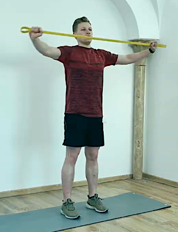Person standing on a yoga mat doing a shoulder stretch with a yellow resistance band, in a room with white walls and wooden floor.