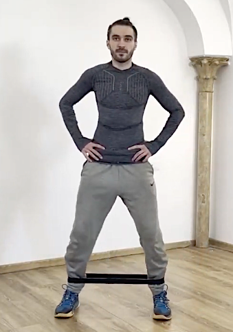 Man standing with hands on hips in a room with white walls and wooden floor, wearing a gray sweater and gray sweatpants, with legs restrained by a black elastic band.