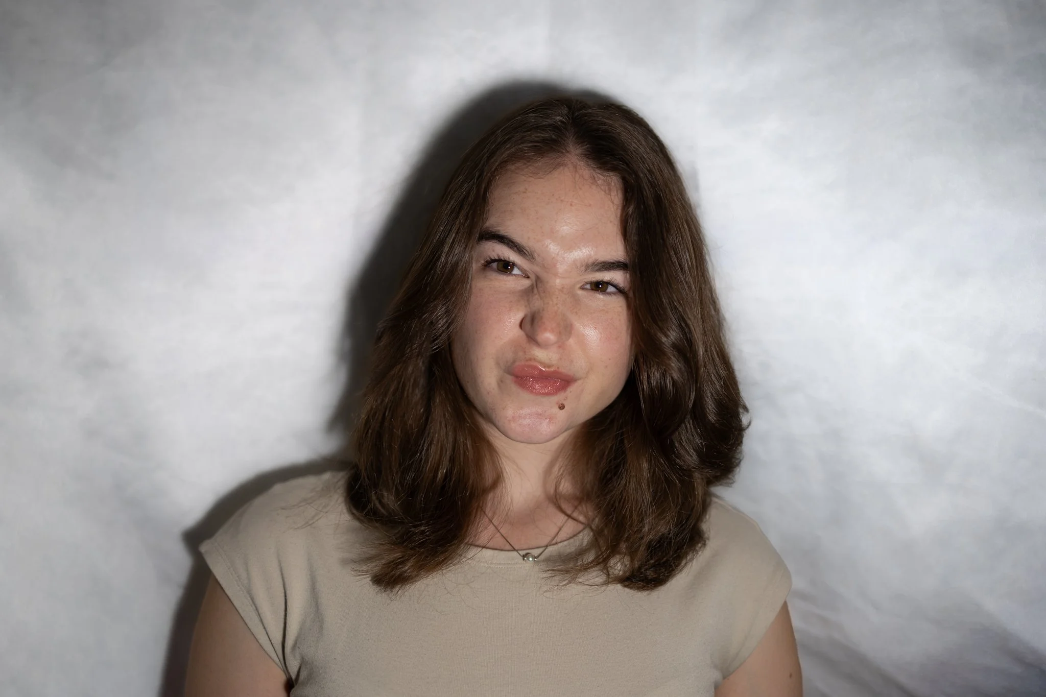 A young woman with light skin, brown wavy hair, and freckles, wearing a beige shirt and a delicate necklace, standing against a plain white wall with shadows cast beside her.