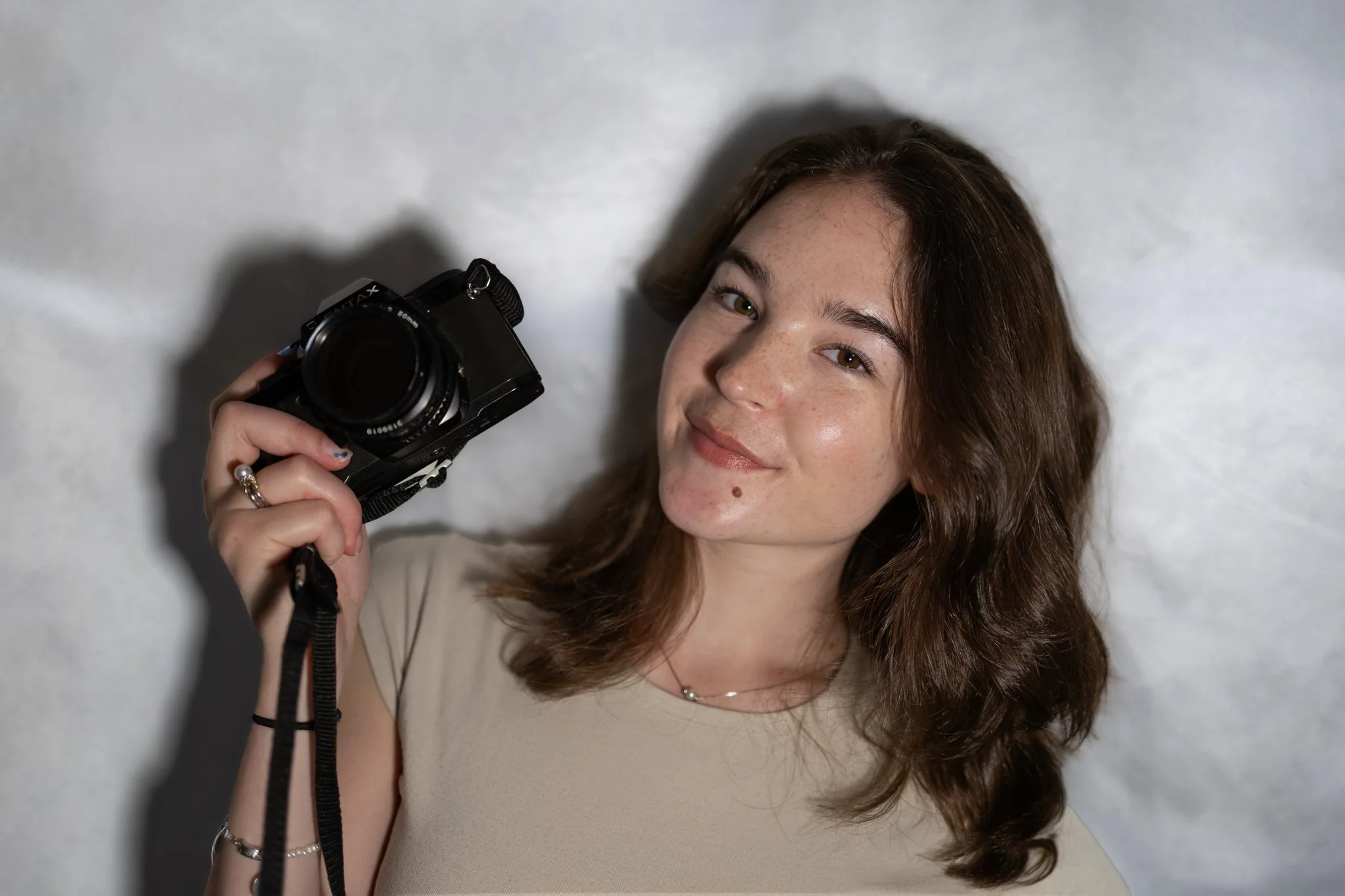 A young woman with long brown hair, freckles, and a small mole on her cheek, smiling while holding a camera in her right hand, against a plain white background.