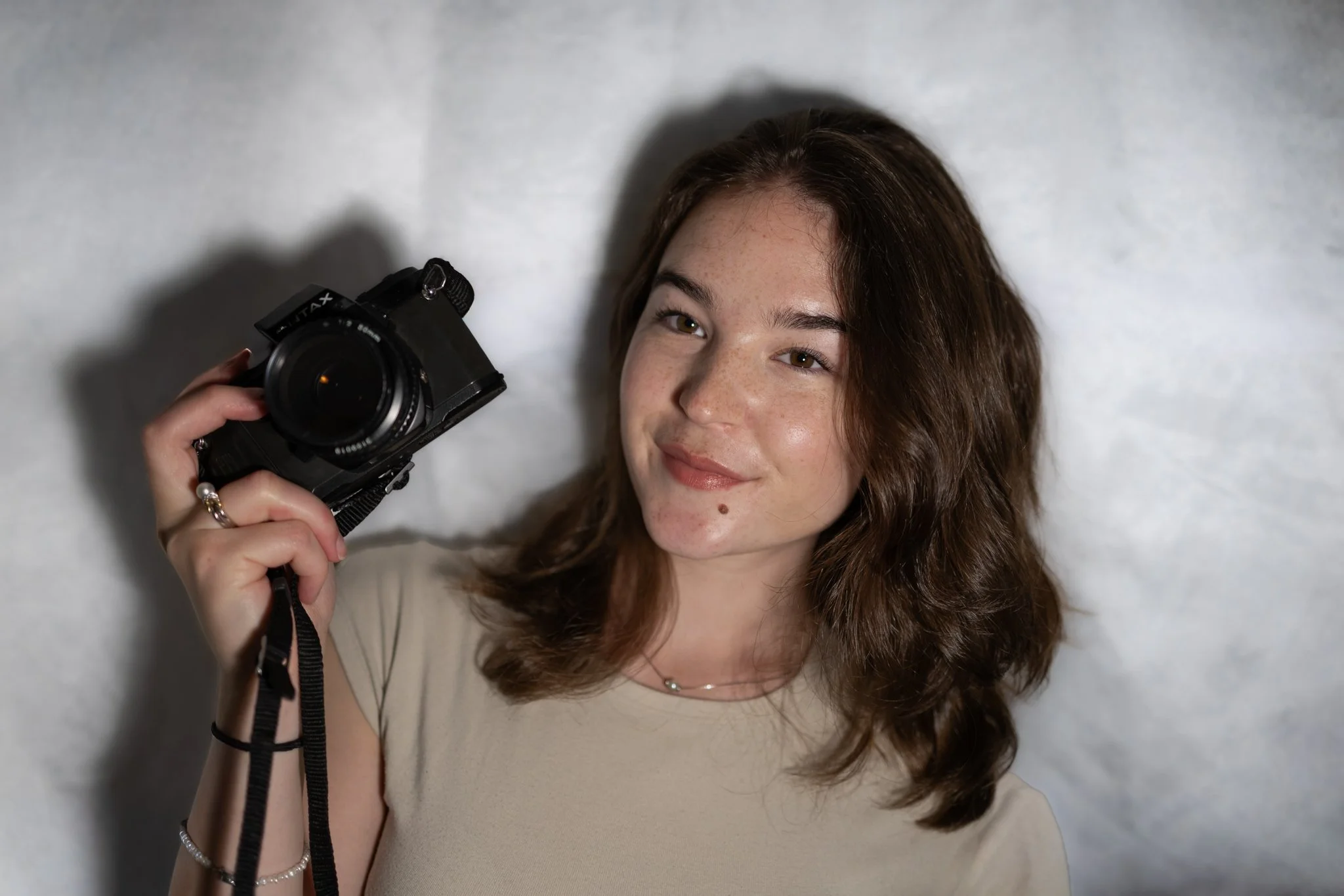 Young woman with brown hair lying on her side on a white surface, holding a black camera in her right hand, smiling slightly.