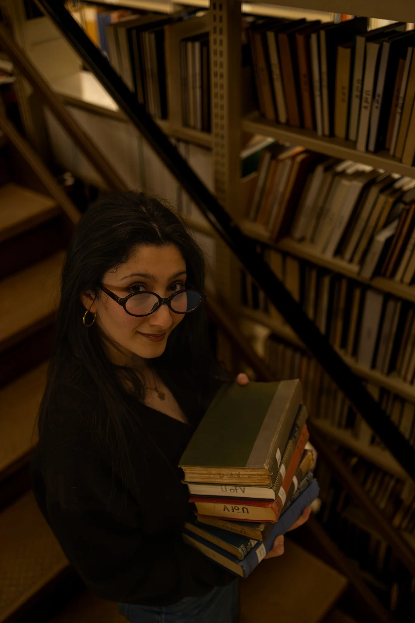 Woman with glasses holding a stack of books in a library with bookshelf filled with books in the background.