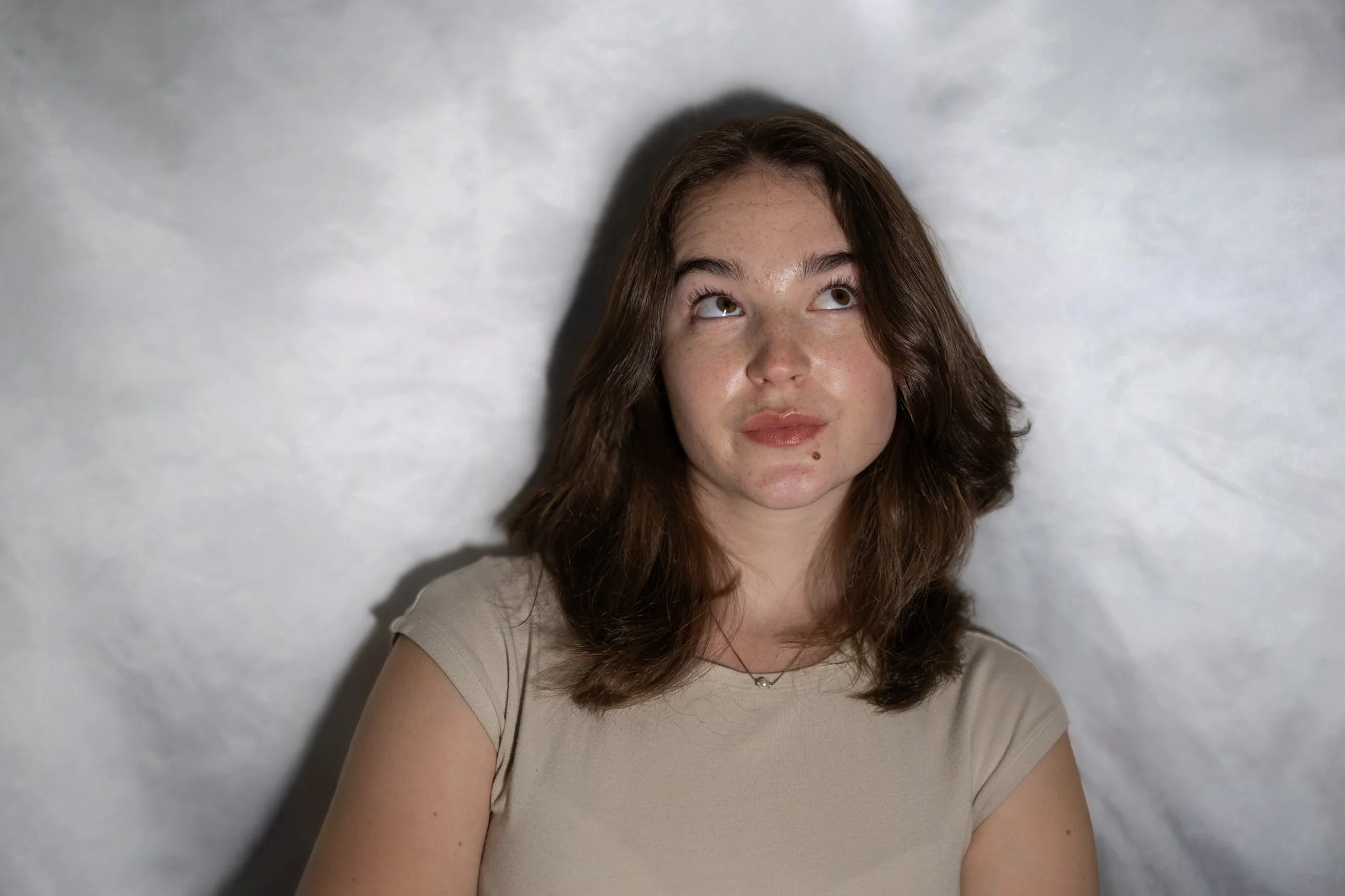 A young woman with brown hair and freckles looking upwards. She wears a beige top and stands against a white textured background.