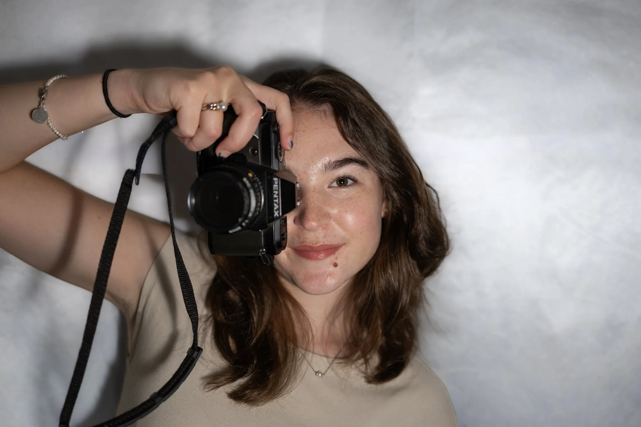 A young woman with brown hair lying down on a light-colored surface, holding a camera up to her eye and looking through the viewfinder.