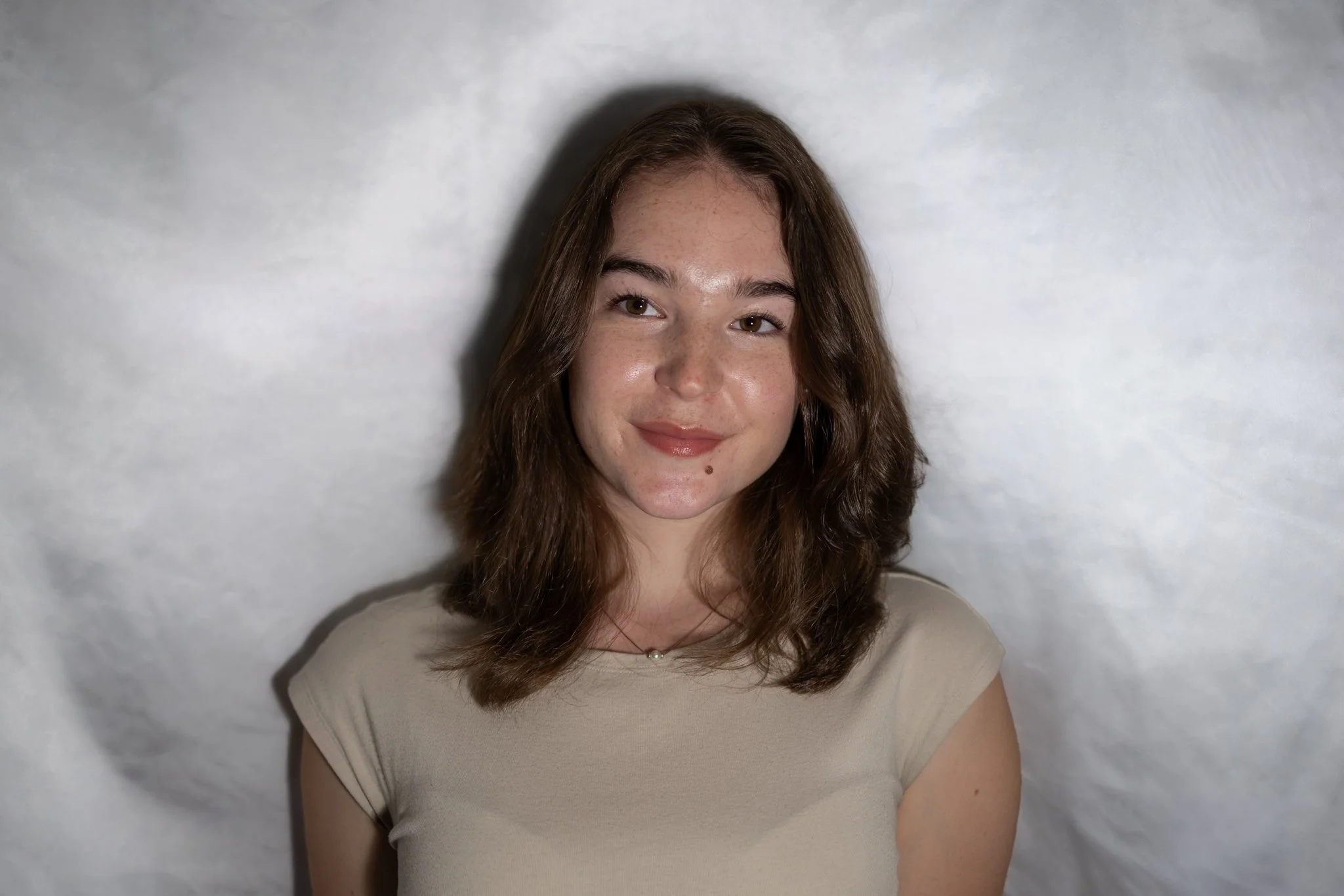 A young woman with light skin, brown hair, and freckles, smiling slightly, sitting against a light-colored background, wearing a beige top.