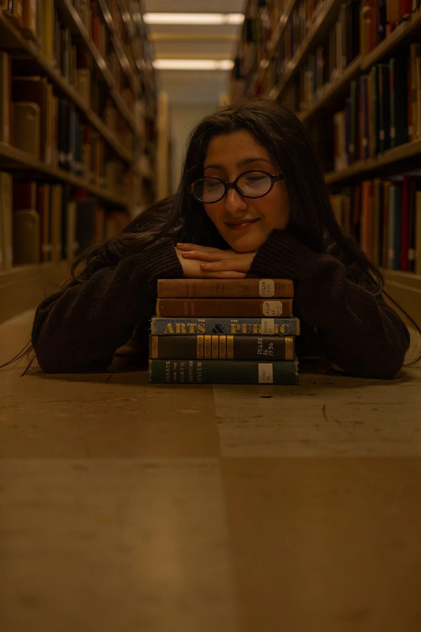 A woman with glasses resting her head on folded hands atop a stack of books in a library aisle.