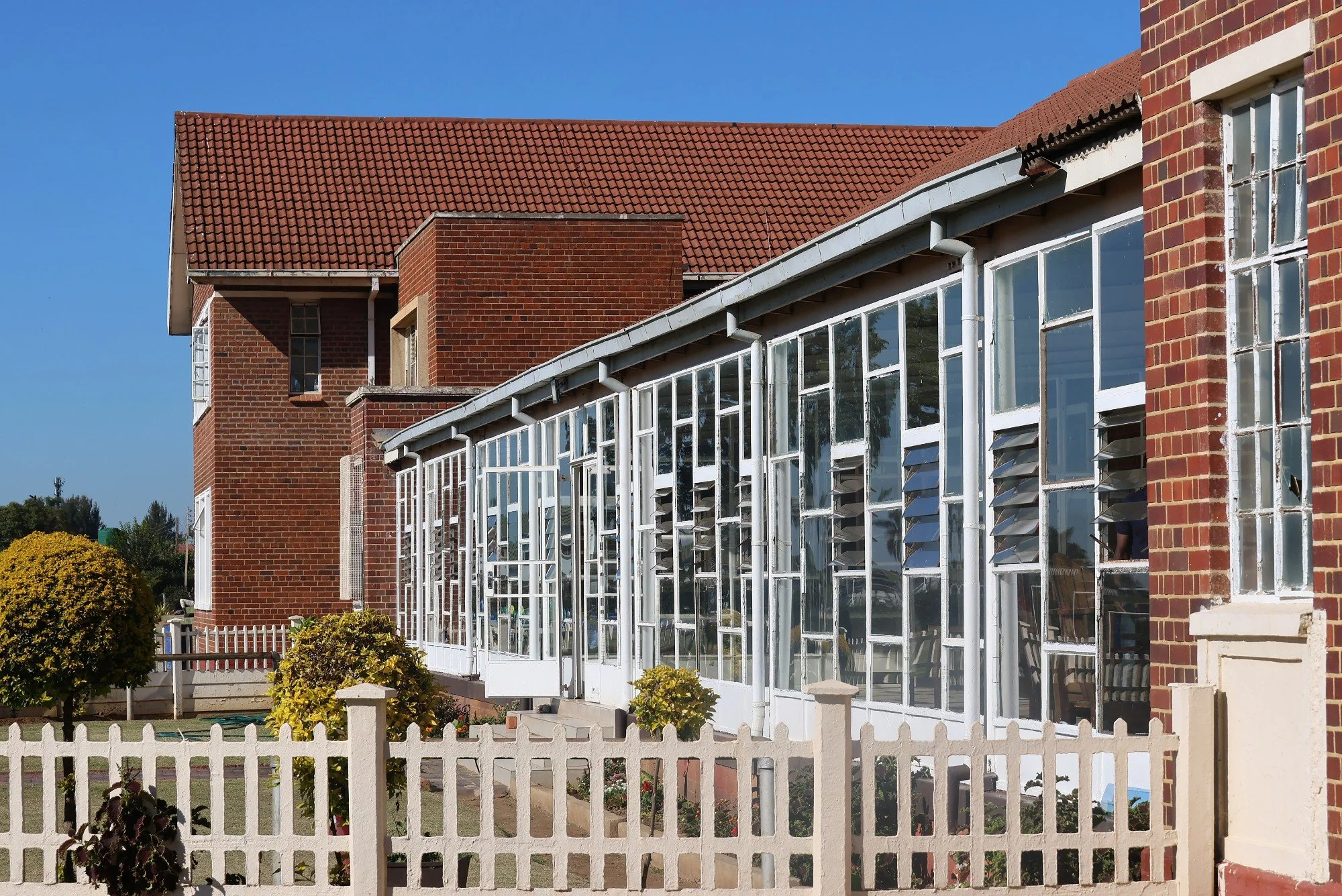 Two-story brick house with a red tile roof and large glass-enclosed porch, white picket fence, green bushes, and clear blue sky.