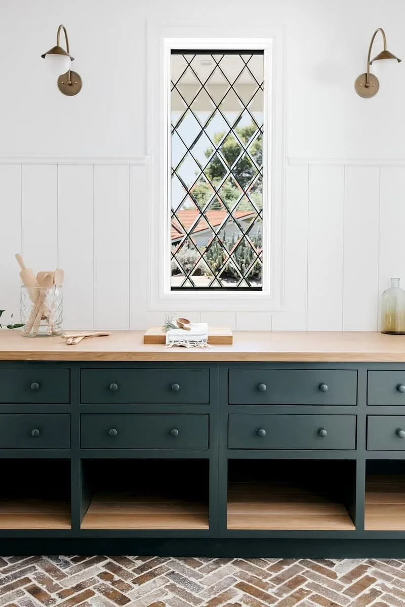 A kitchen with a green cabinet, wooden countertop, and a Pella Reserve window with diamond-patterned window panes. The wall behind the counter has white paneling, and there are wall sconces on either side of the window.