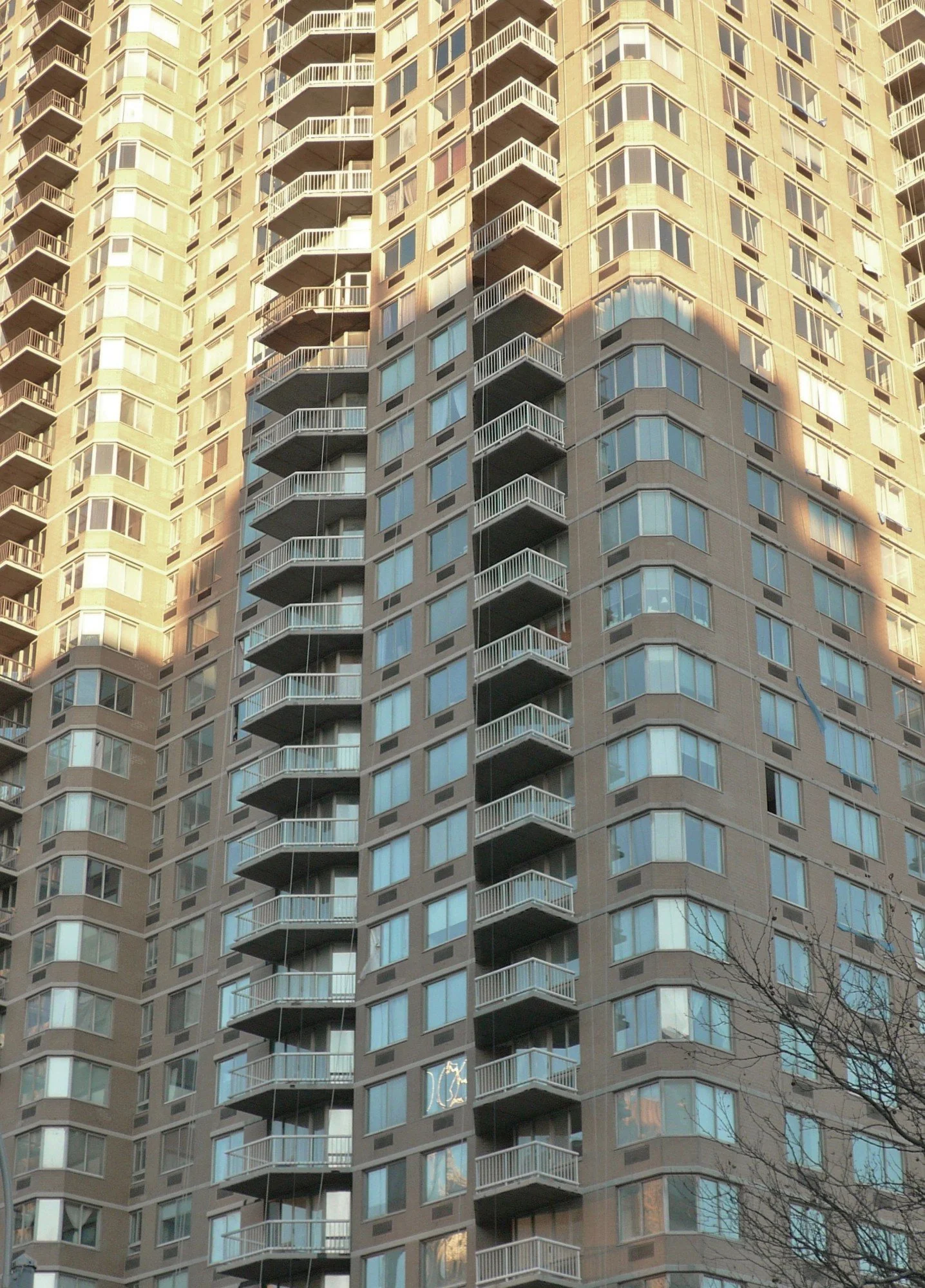 A tall residential apartment building with multiple balconies and large windows. There are reflections of the sky and other buildings in the windows. A tree is visible at the bottom right corner.