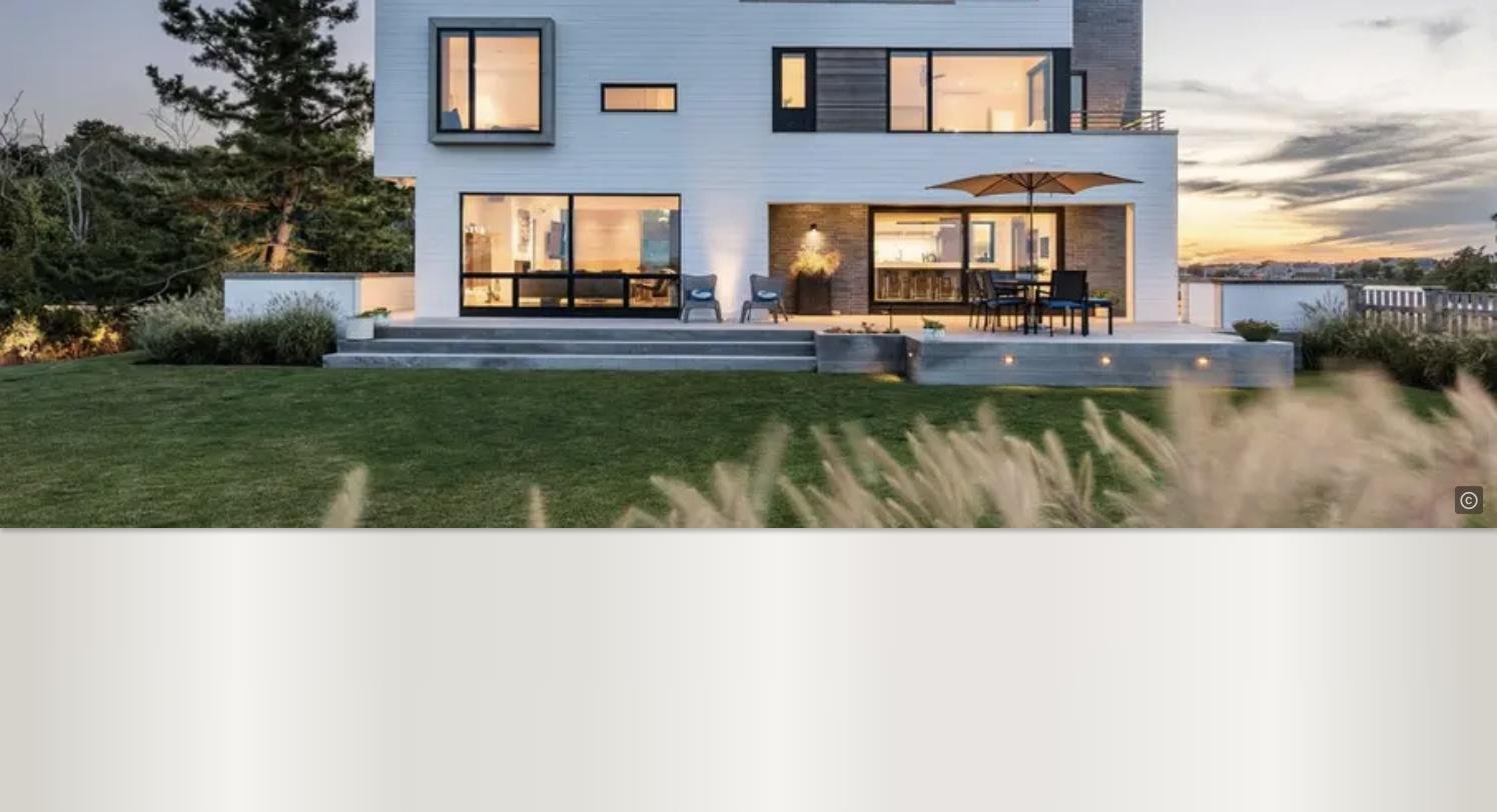 Modern house with large glass windows, outdoor patio with chairs, table, and umbrella, green lawn, trees in the background, and sunset sky.