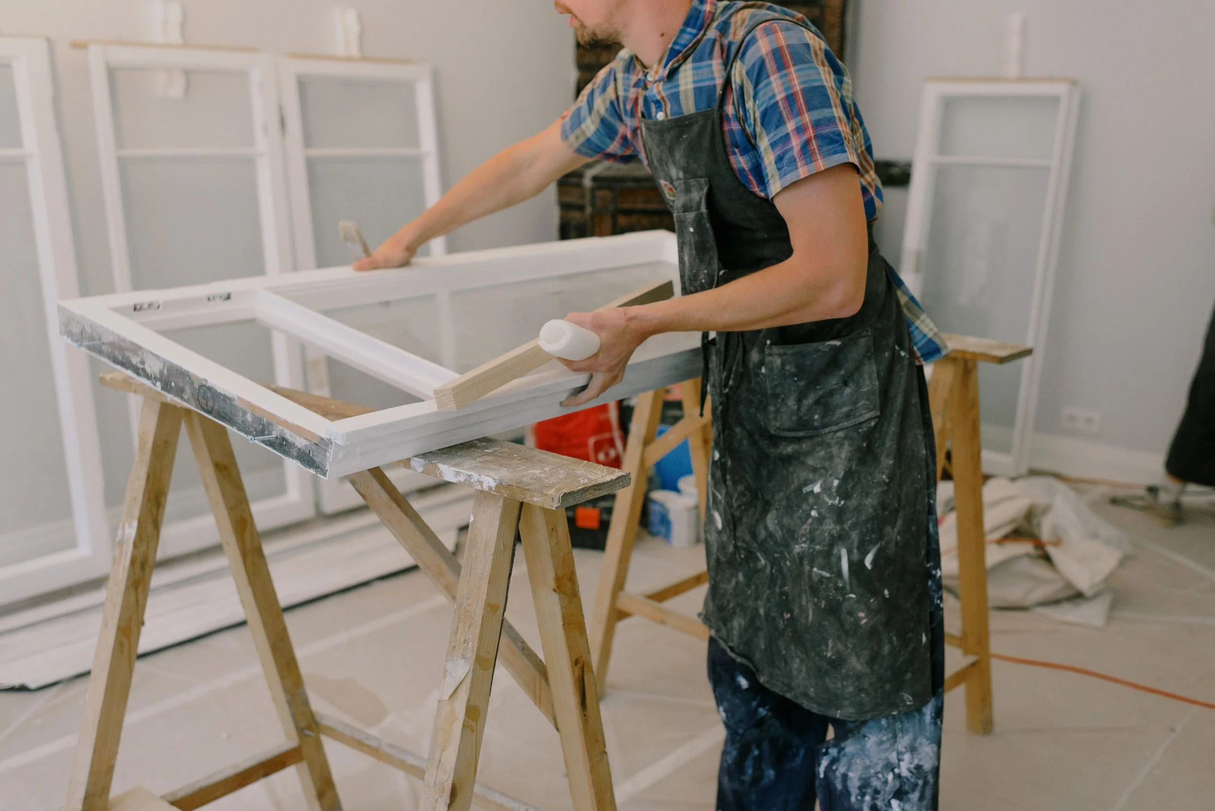 A man wearing a plaid shirt and black apron working on a woodworking project at a workshop, using a brush on a white-painted window frame on a wooden sawhorse.