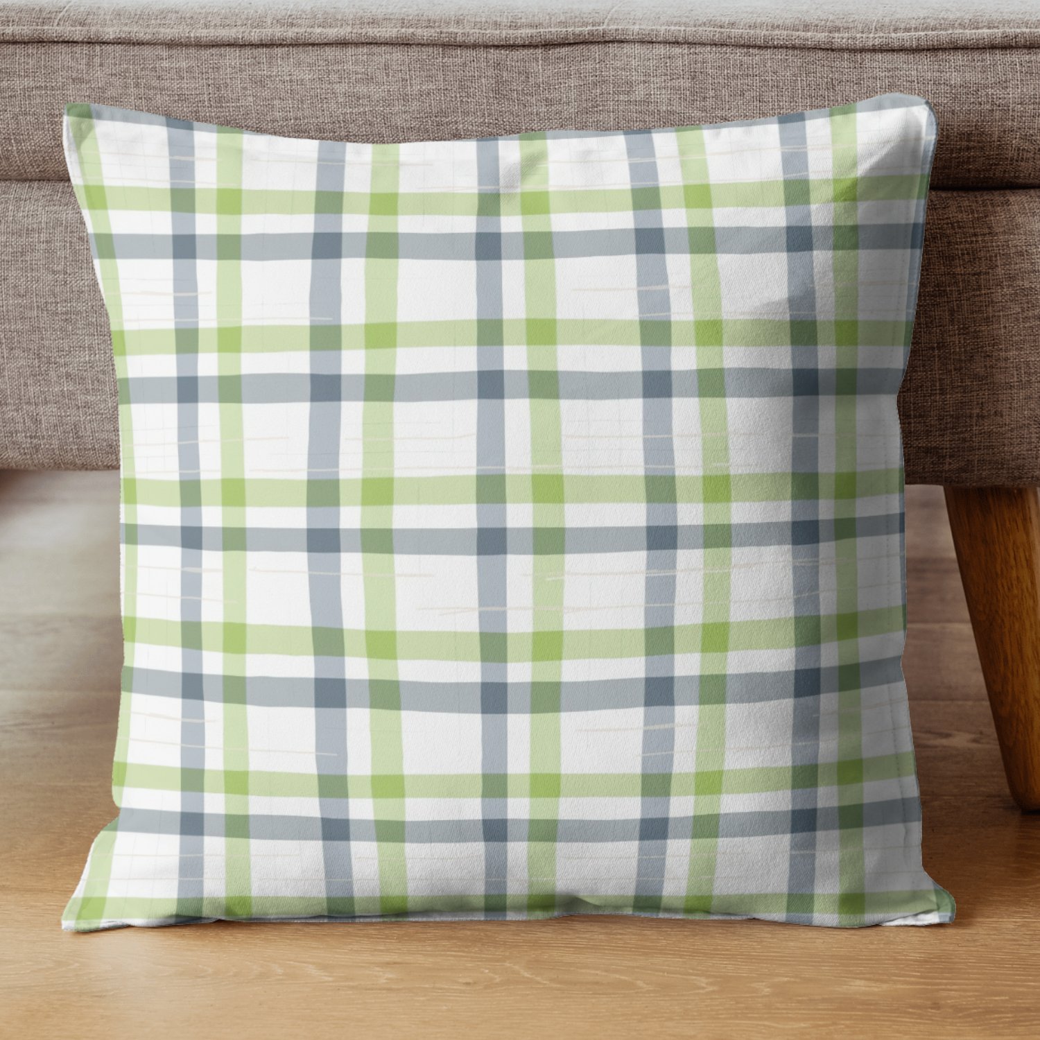 A plaid decorative pillow with green, gray, and white colors on a wooden floor in front of a brown sofa.