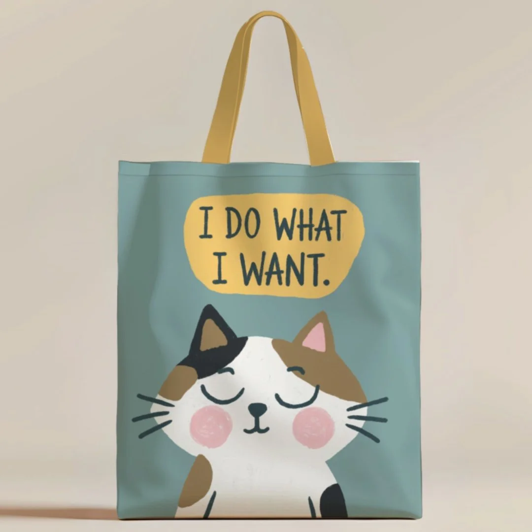 Light blue tote bag with yellow handles, featuring a cartoon cat with closed eyes and pink cheeks. Above the cat, text reads, "I DO WHAT I WANT."