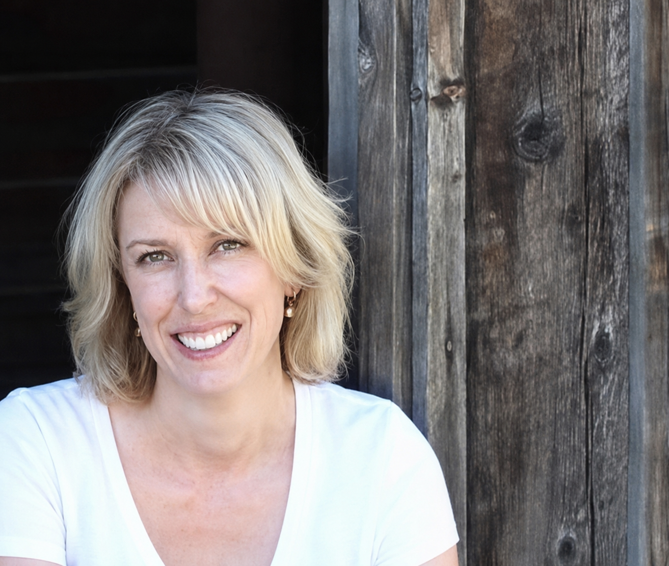 Cindy Craig, the artist of Jolly Little Prints. She has Blonde hair, smiling, wearing a white shirt and earrings, sitting outdoors with a wooden wall in the background.