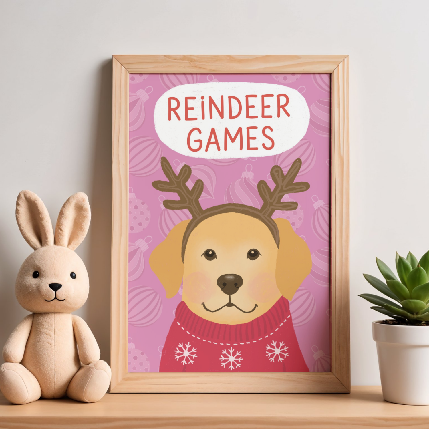 Decorative framed poster with a reindeer wearing a red sweater with snowflakes, and the text "Reindeer Games" in red letters on a pink background with ornament patterns. To the left, a plush bunny toy, and to the right, a potted succulent plant.